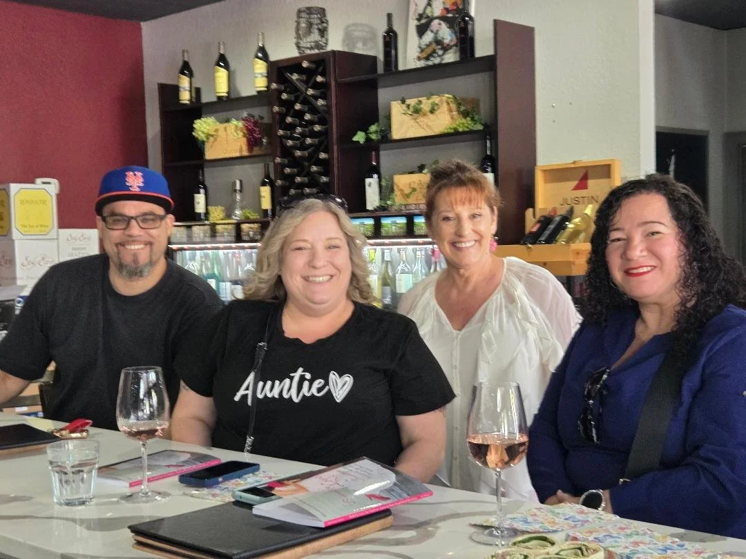 Four people sitting at a table in a restaurant or bar, smiling at the camera. There are two women and one man facing the camera and one woman facing slightly to the right. The table has wine glasses, a menu, and a phone. Behind them are shelves with wine bottles and decorations.