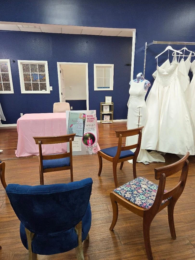 Room setup with chairs, wedding dresses on a rack, and a table with sign-up materials, indicating a bridal gown shopping or fitting event.