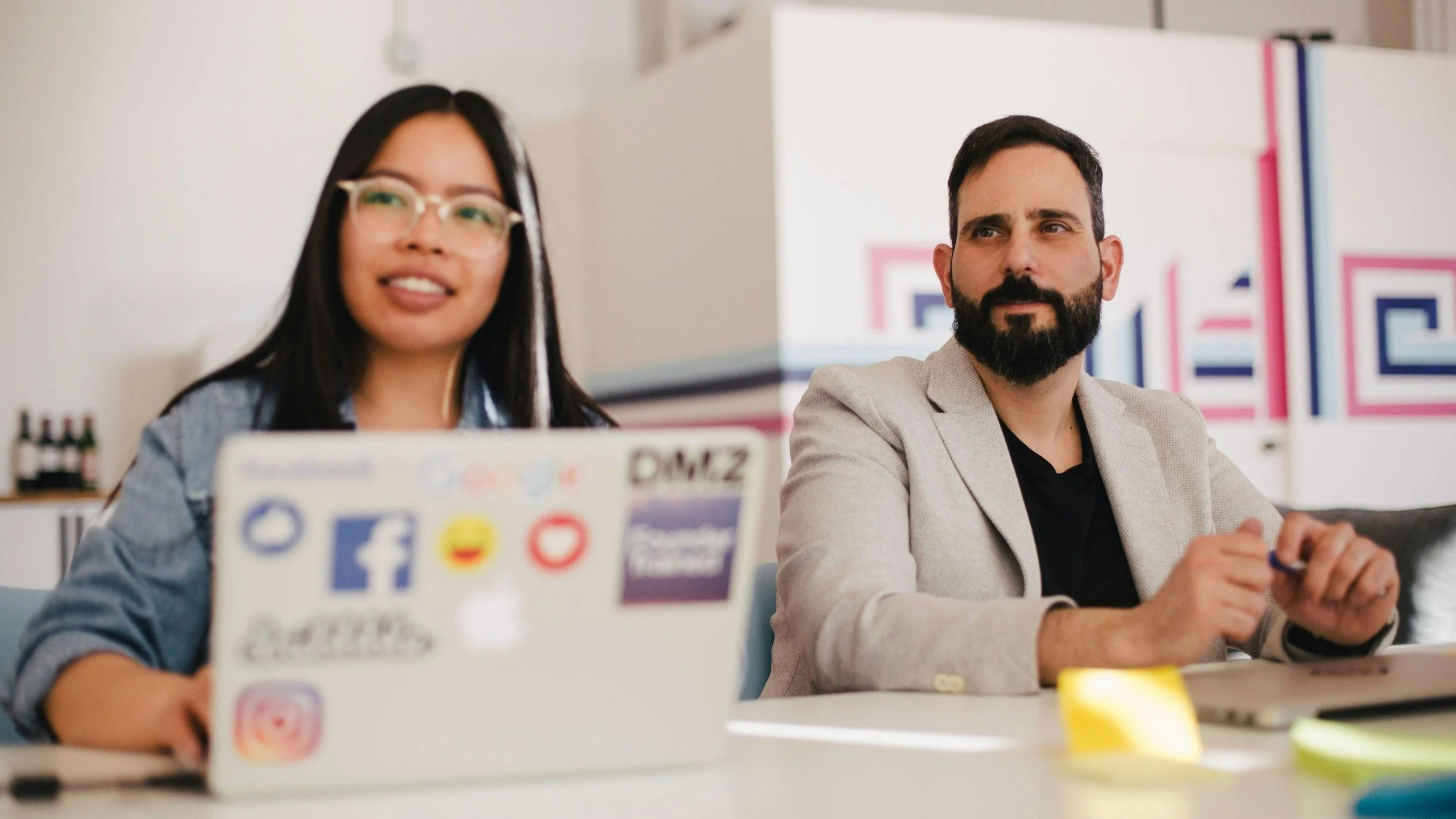 Young woman and middle-aged man sitting in a meeting room, looking up attentively with polite but slightly skeptical expressions.
