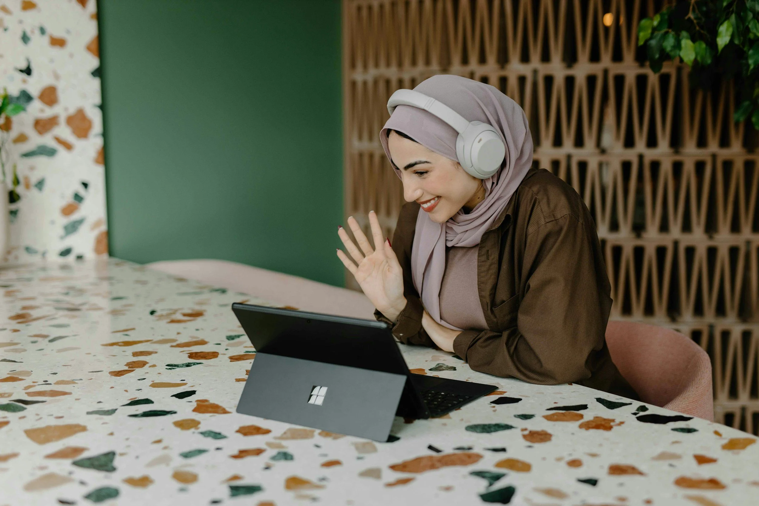 Woman wearing headphones and a head scarf, smiling and waving during a video call on her tablet — confident, connected, and ready to engage.