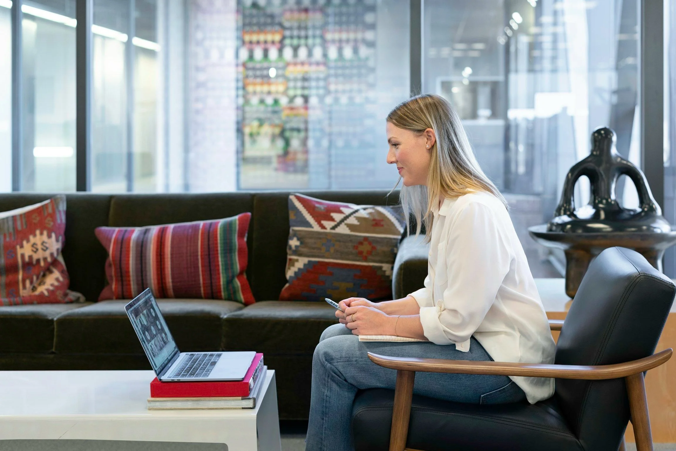 A woman sitting on a black chair in a modern office lounge, looking at her phone, with an open laptop and colorful pillows behind her.