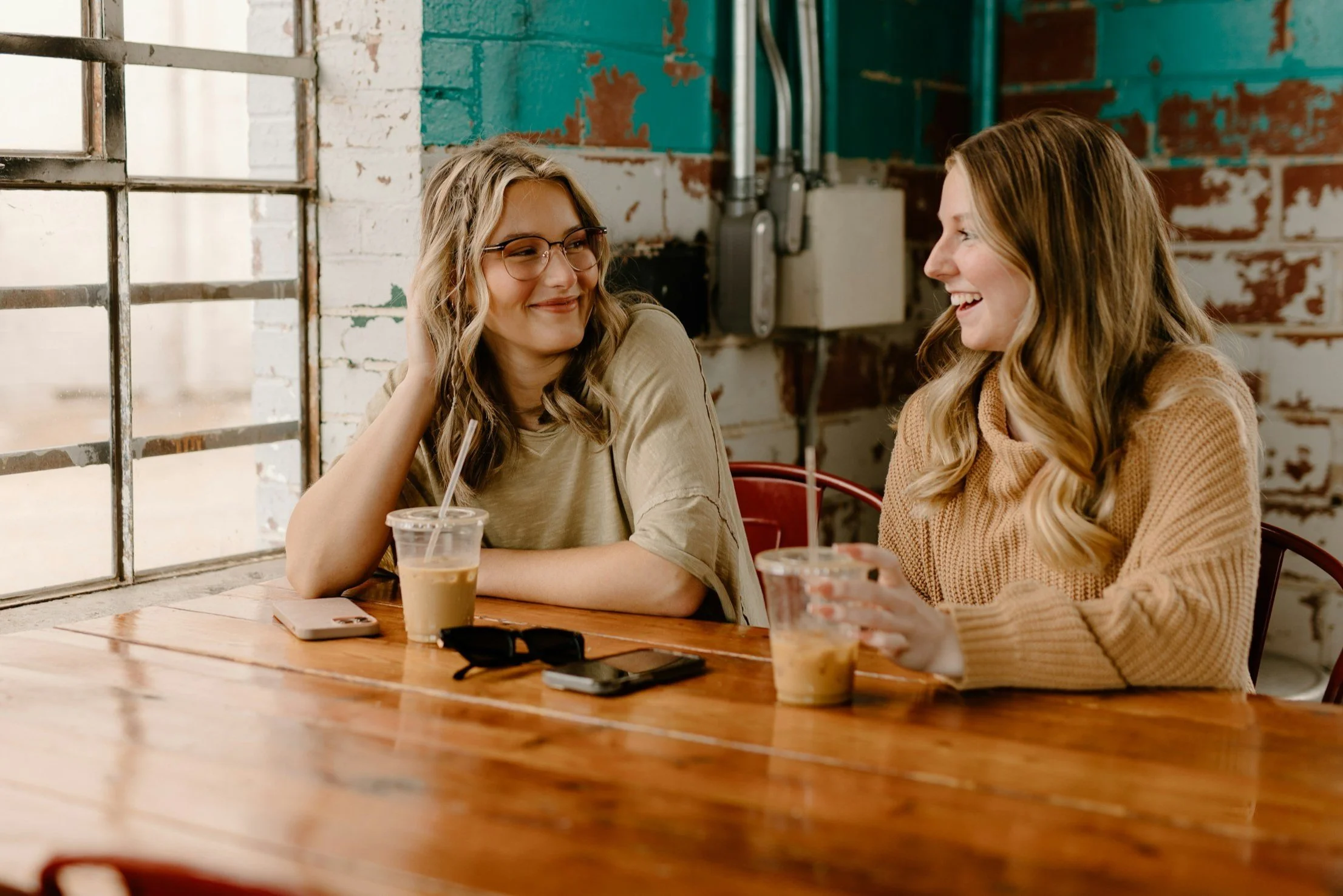 Two women sitting at a wooden table in a coffee shop, smiling and having a conversation, with iced coffee drinks and phones on the table.