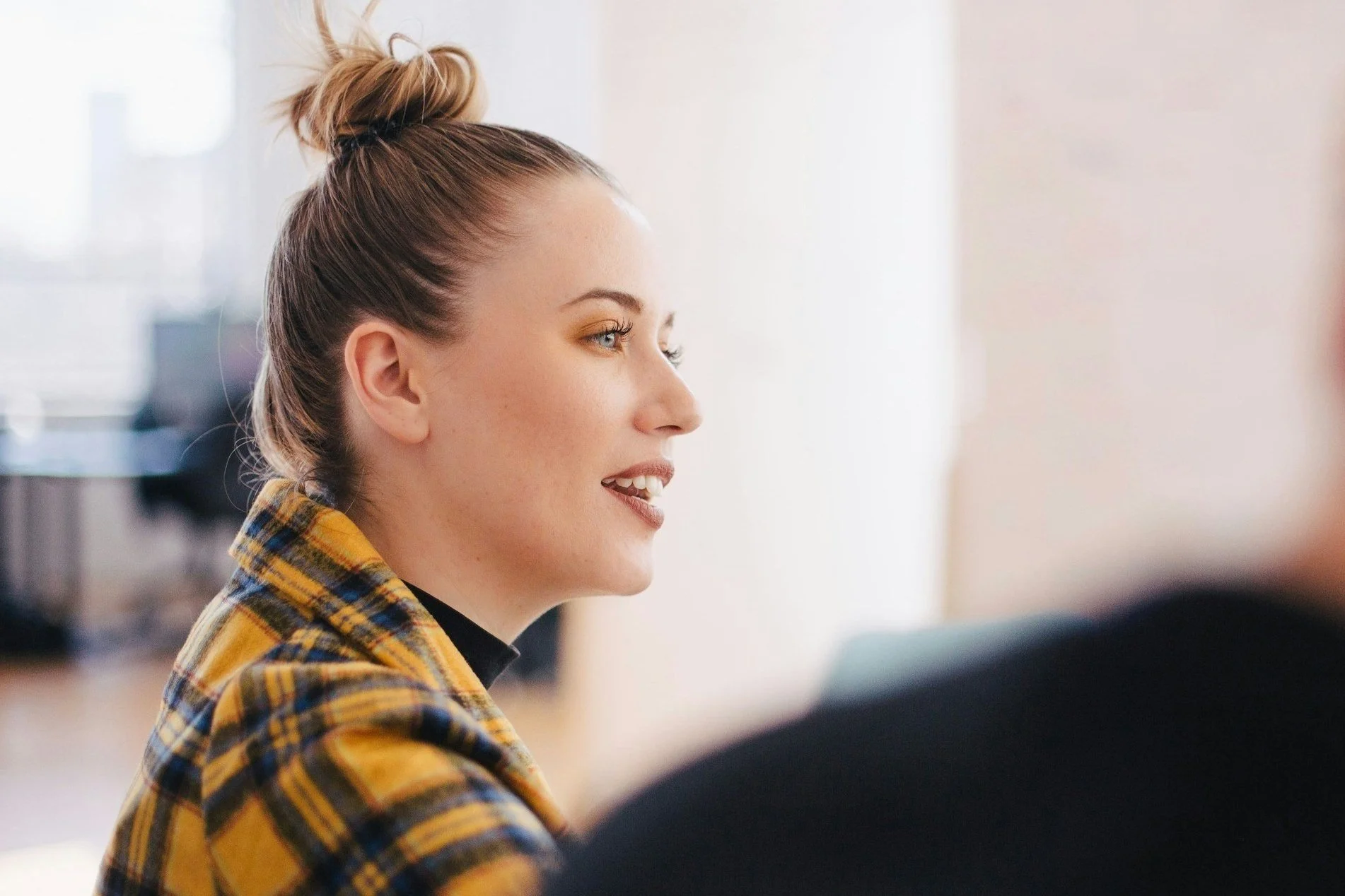 Confident woman in a plaid jacket speaking in a meeting room, captured mid-sentence as she shares a clear, composed message.