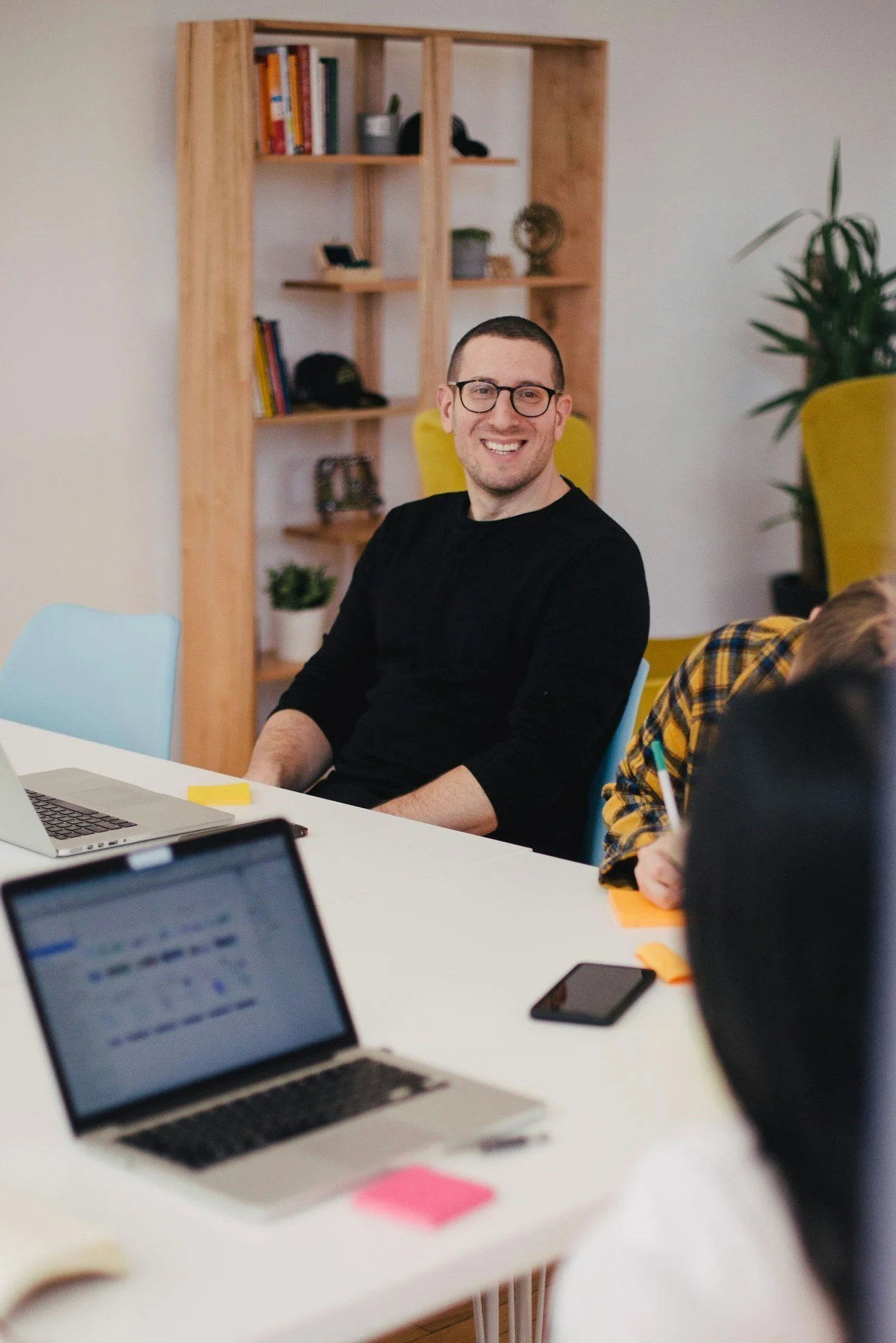 A man with glasses and a black shirt sitting at a table, smiling. There are laptops and a phone on the table. Shelves with books and decorative items are in the background.