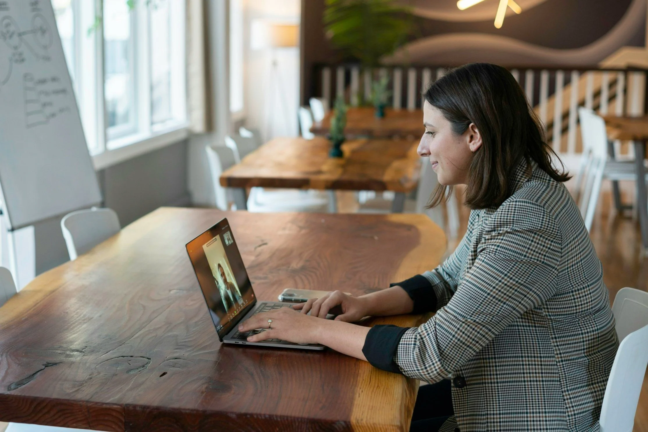 Woman sitting at a wooden table, engaged in a video call on her laptop, in a bright, modern room with large windows and white chairs.