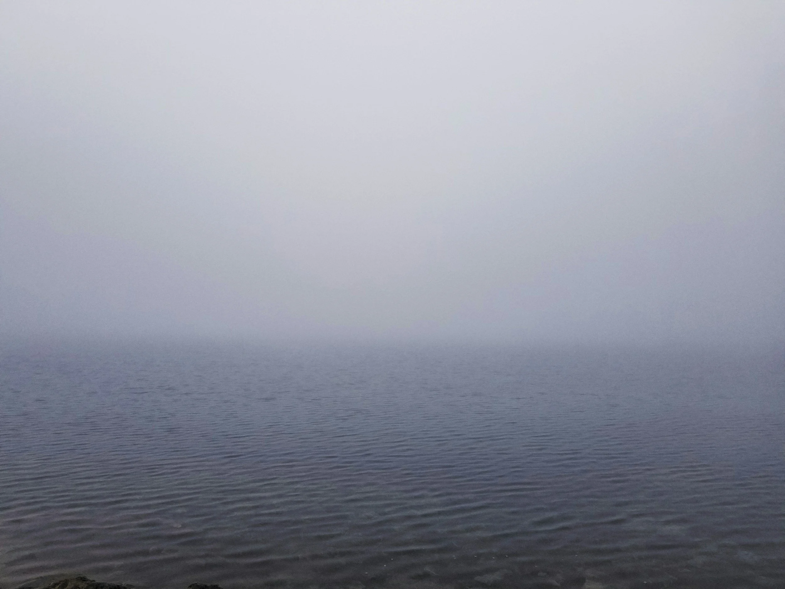 A foggy body of water with a fog-covered sky, and mountains barely visible in the background.