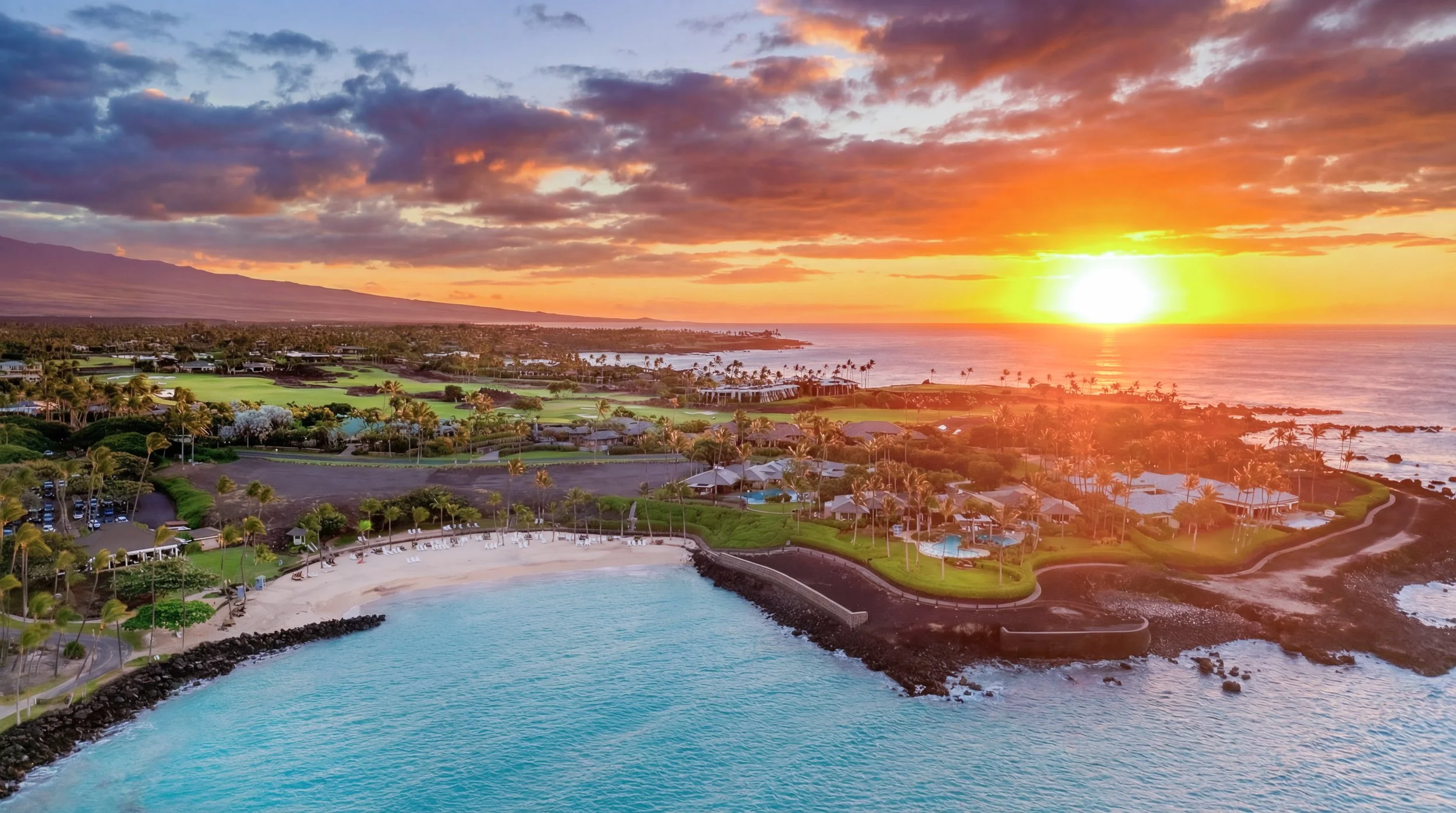 Aerial view of a coastal resort during sunset with a beach, swimming pools, palm trees, and a scenic ocean shoreline.