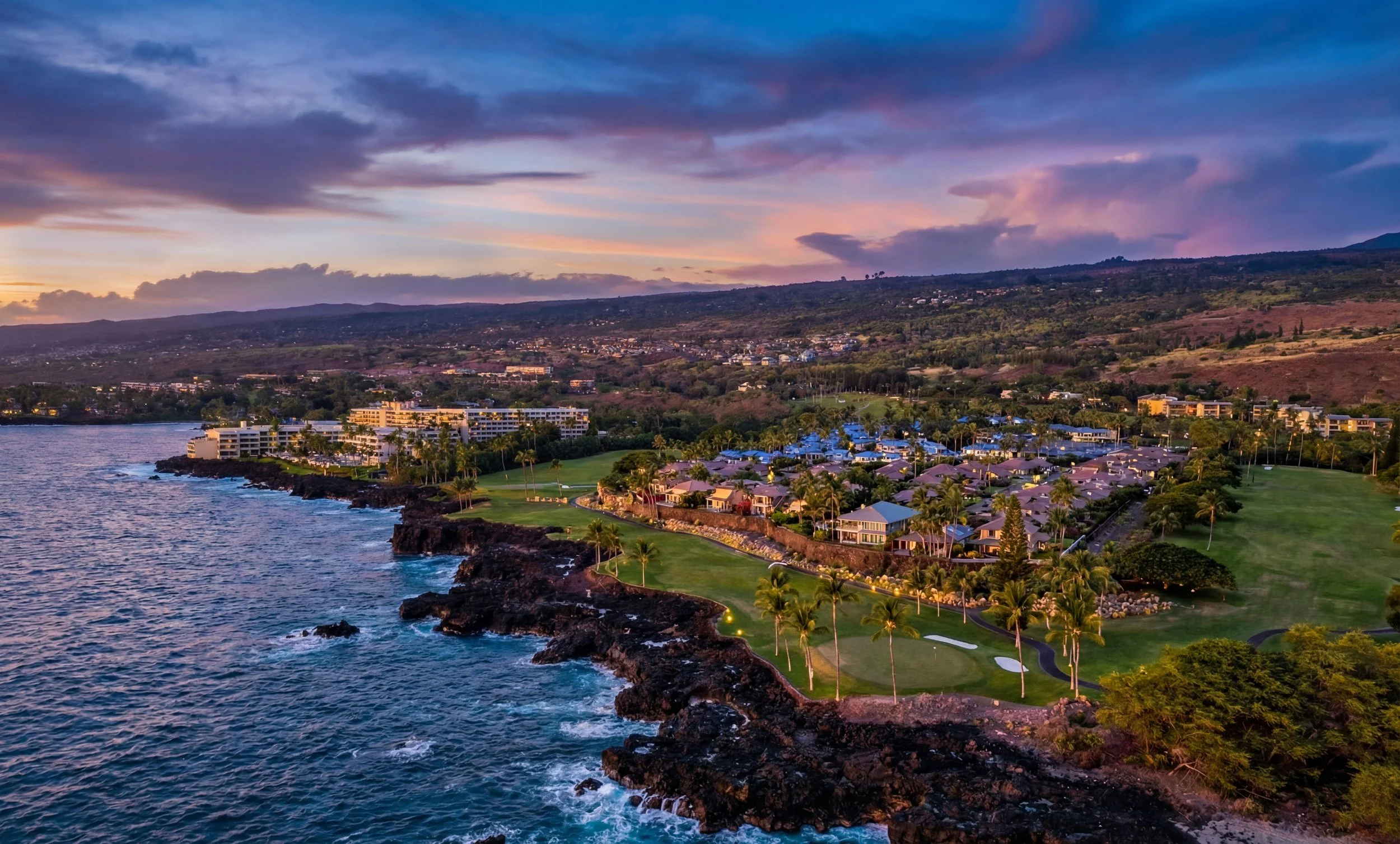 A coastal landscape at sunset with a golf course, residential buildings, and a hillside in the background.