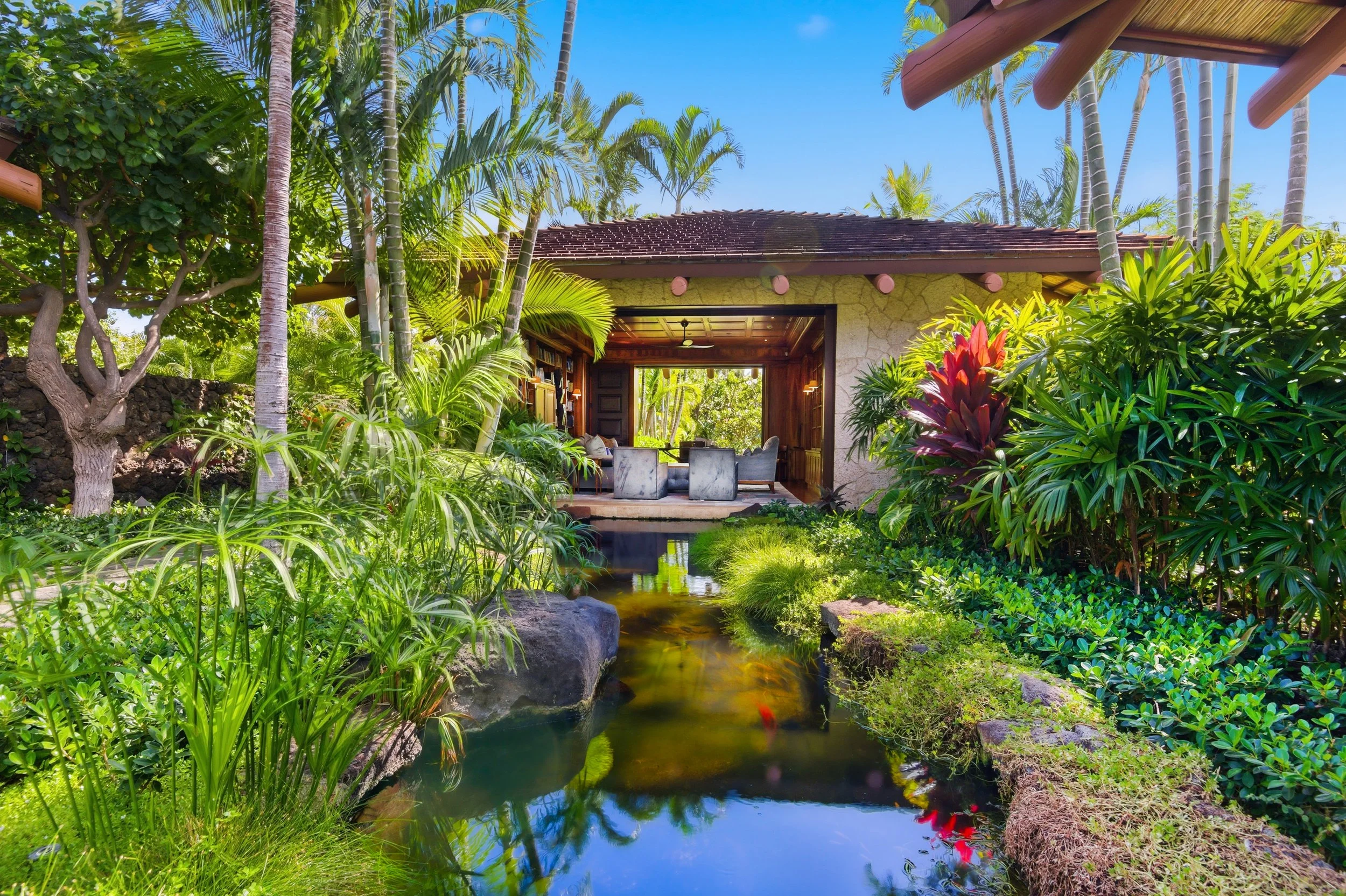 A lush tropical garden with a small pond, various green plants, trees, and a seating area in front of a house with a brown roof and open wall in the background.