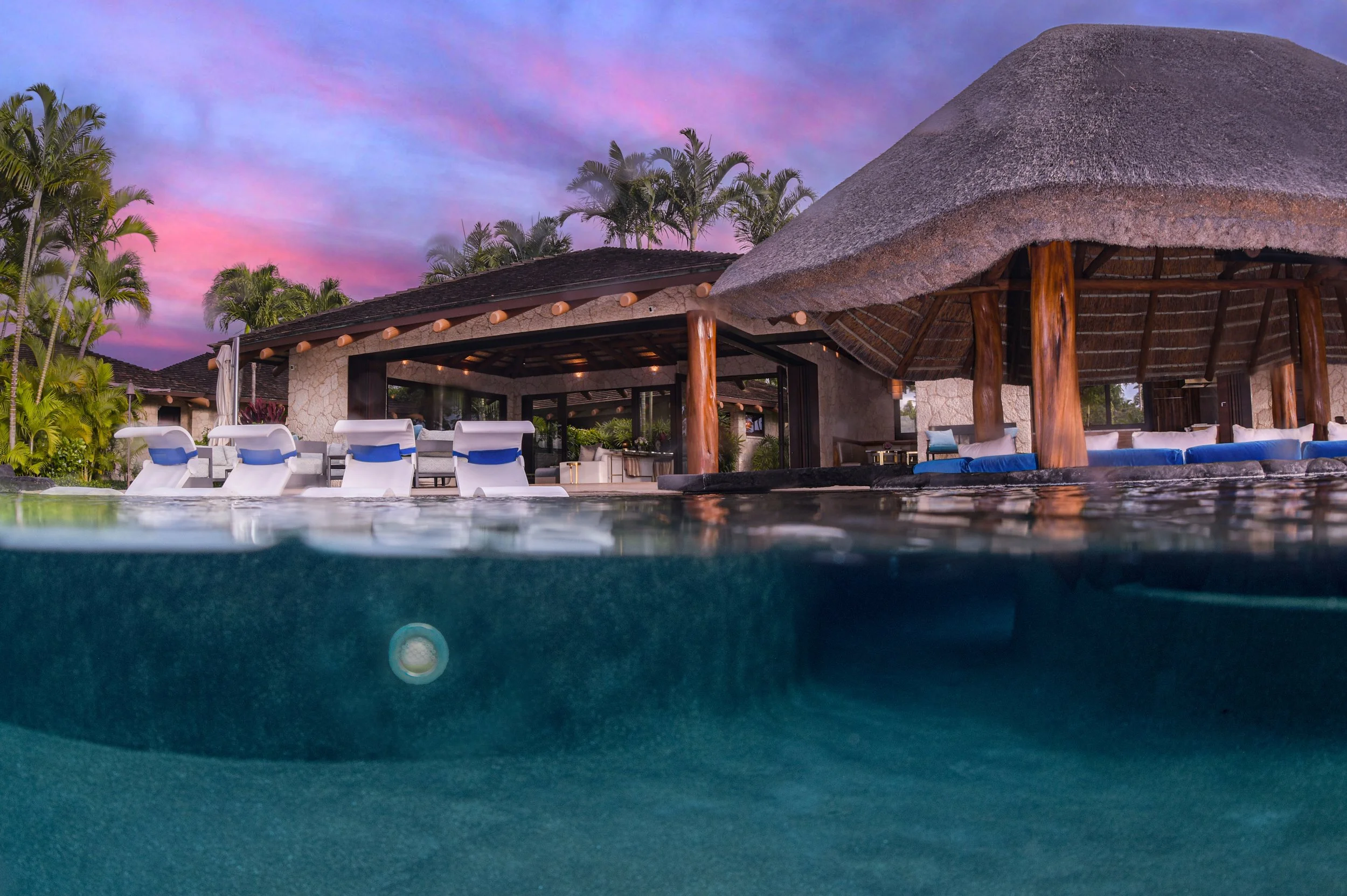 Overwater pool with mountain-style thatched roof structure, lounge chairs, and tropical palm trees at sunset.