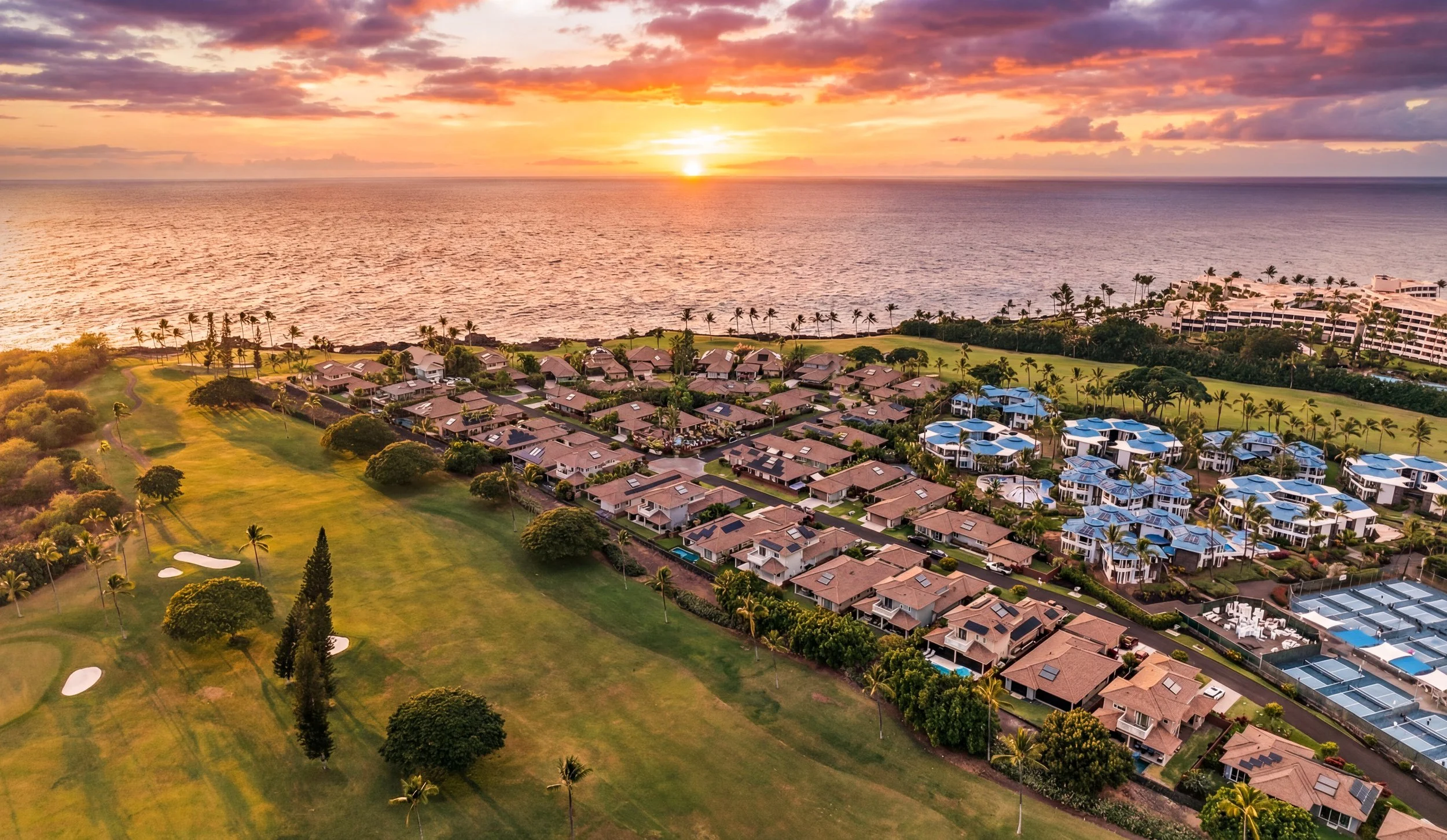 Aerial view of a coastal neighborhood at sunset with ocean, palm trees, houses, golf course, and buildings