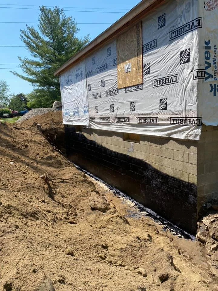 Side of a house under construction or renovation, with house wrap insulation and black waterproofing at the base, and dirt excavation along the foundation.