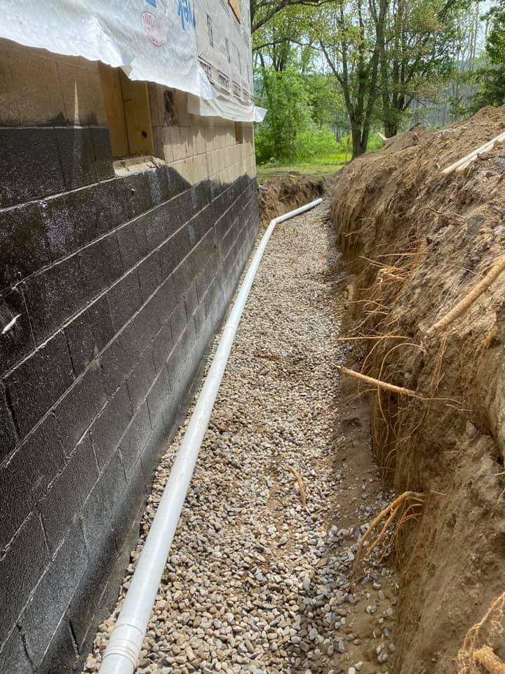 Construction site with black foundation wall, gravel pathway, white drainage pipe, and dirt embankment alongside a house with fiberglass insulation visible at the top.