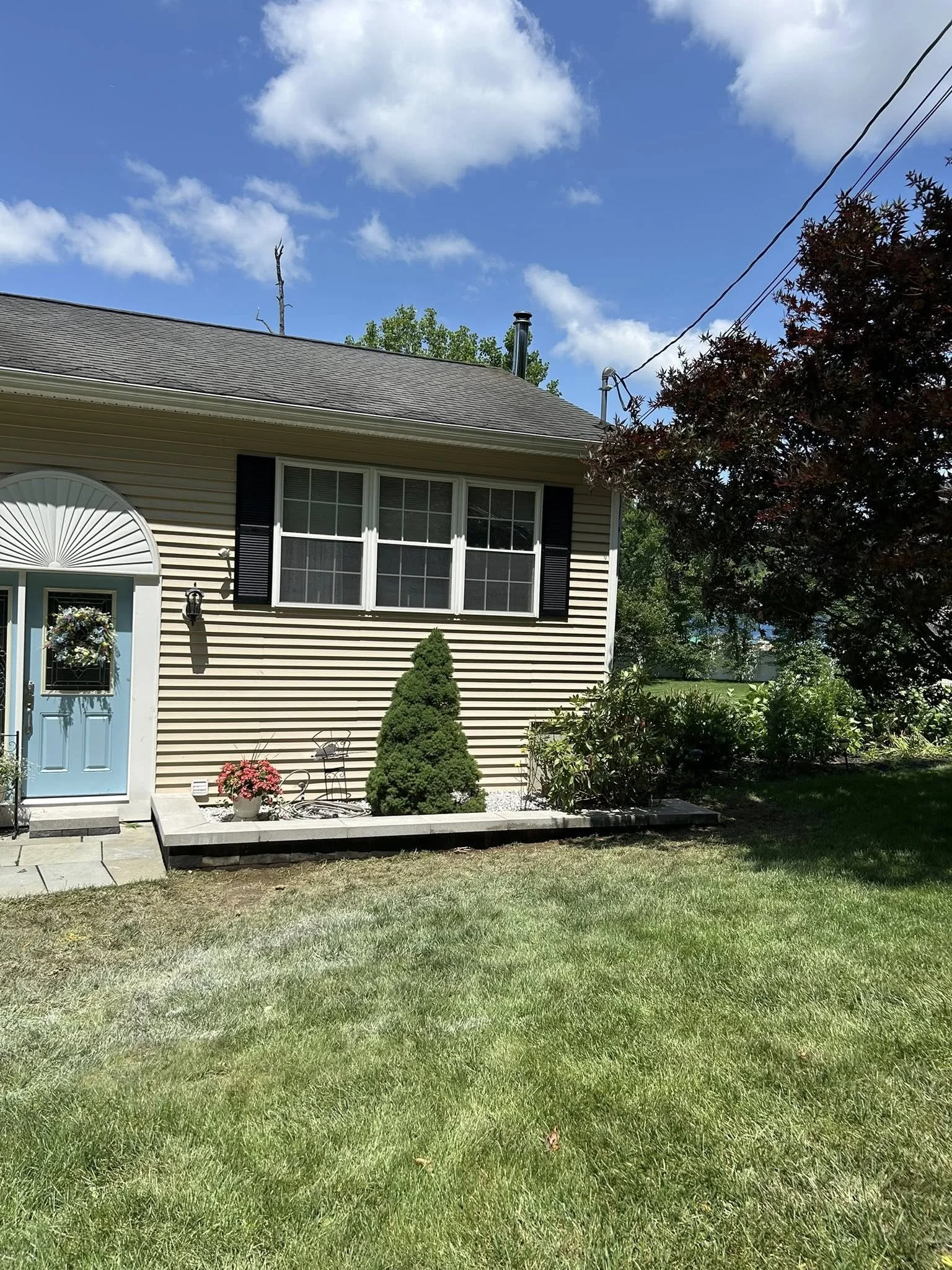 Front yard of a house with a lawn, a small garden bed with plants and a trimmed bush, and a beige house with a blue door, window shutters, and a large window. There are clouds and blue sky overhead.