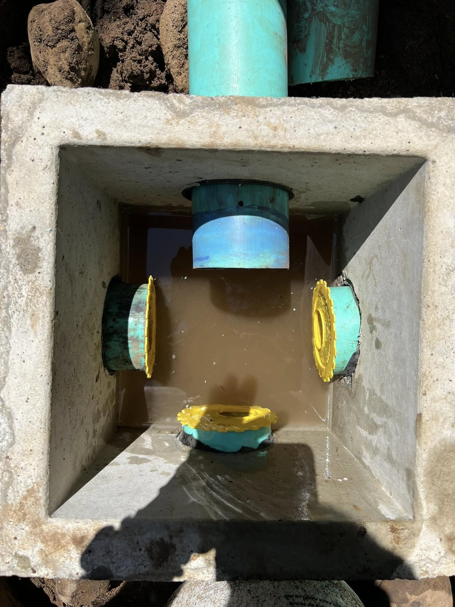 Underground equal flow  distribution box with four pipes and some water filled inside, surrounded by dirt.
