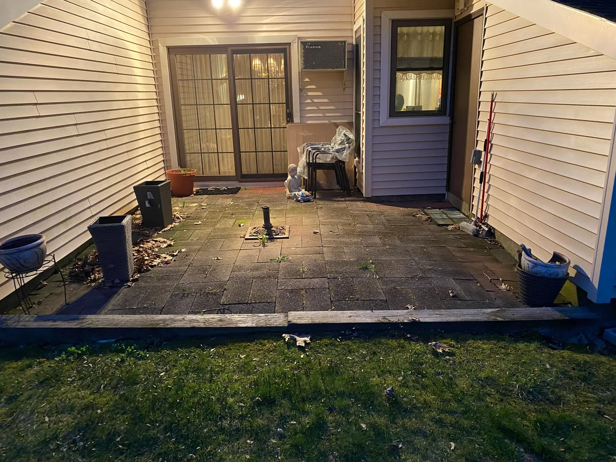 A small outdoor patio enclosed by white siding walls, with a sliding glass door, a small window, and plants in pots. The patio has paver stones, some dirt, debris, and condensation on the glass door. There are chairs stacked in the corner, garden too