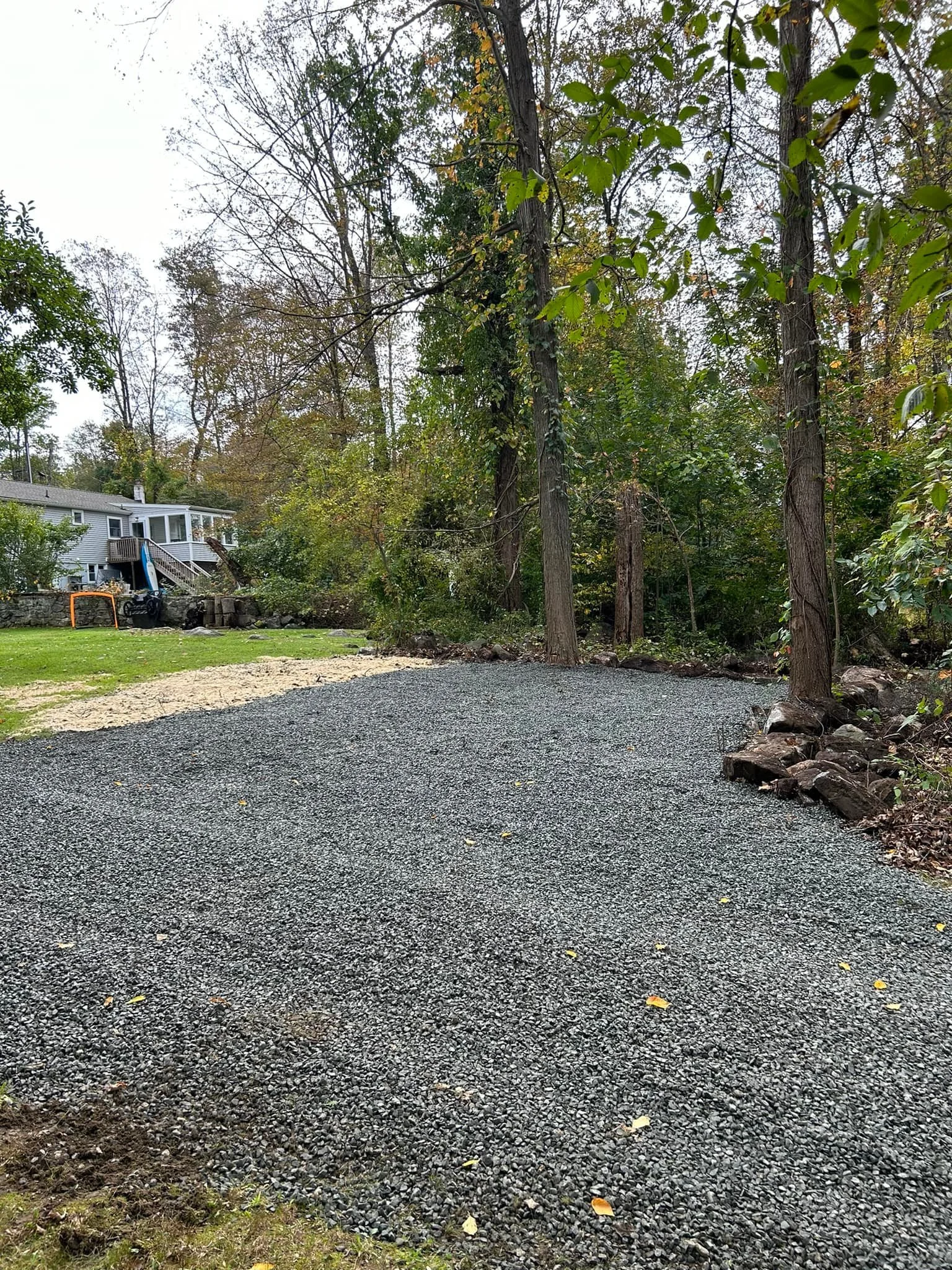 A backyard with a gravelly area next to a grassy lawn and trees, with a house in the background.
