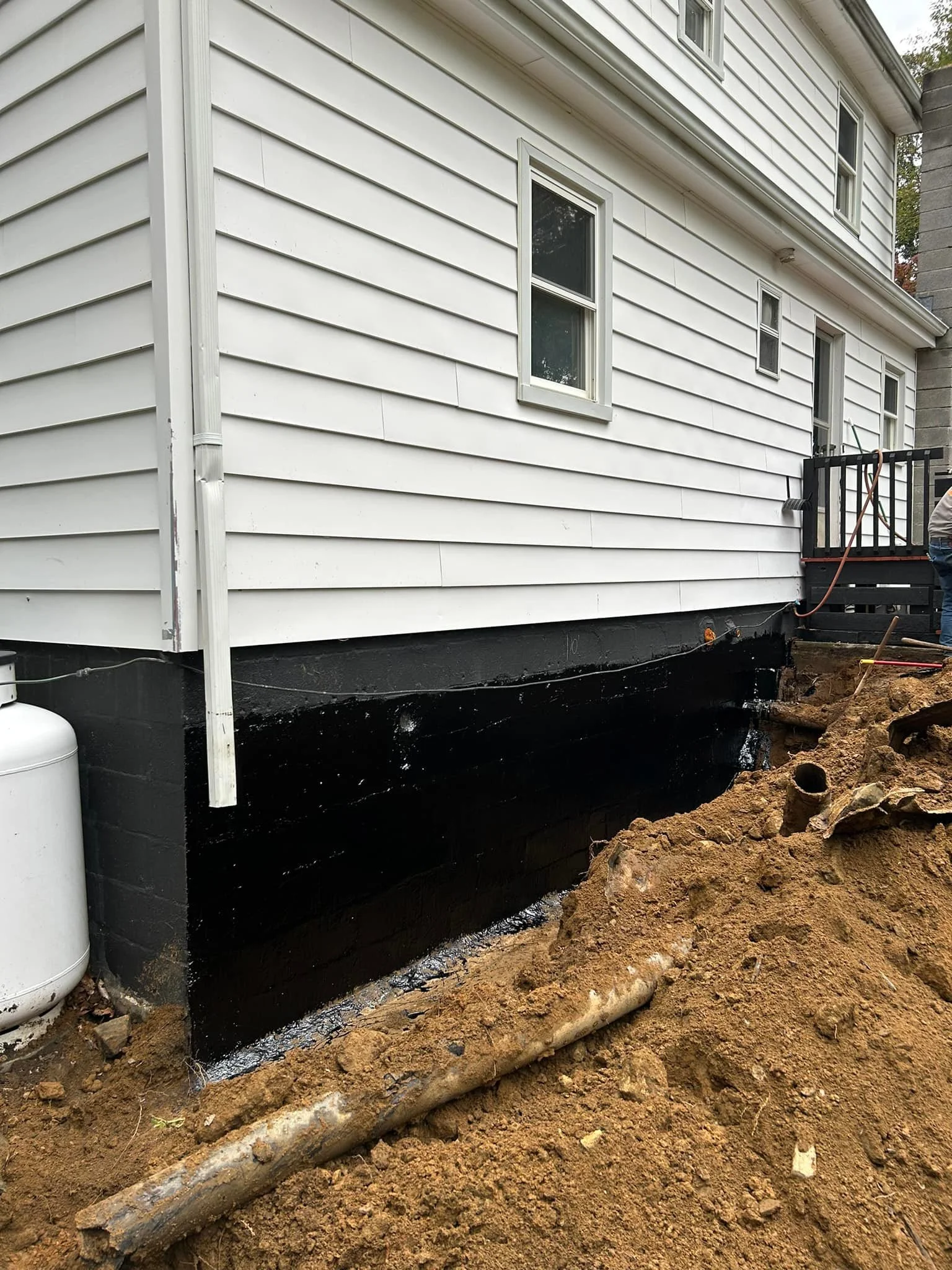 Back of a house with white siding, partially excavated with black waterproofing on the foundation.