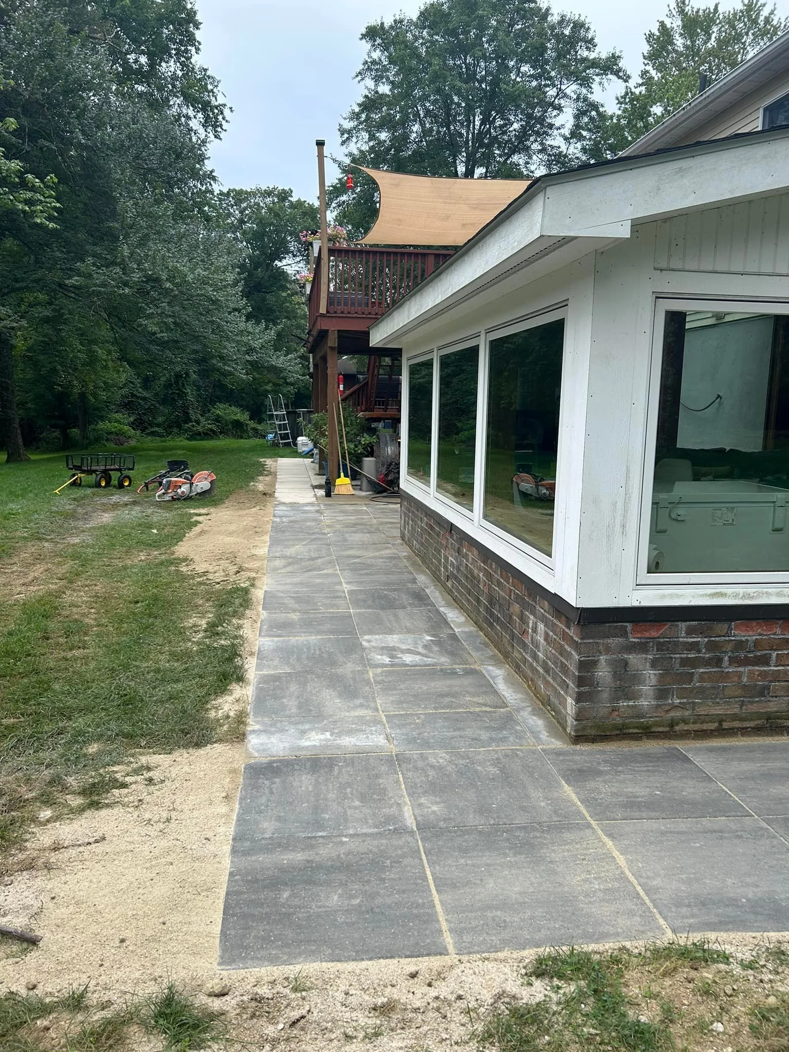 Backyard with a completed stone patio adjacent to a white house with large windows, and a wooden deck.