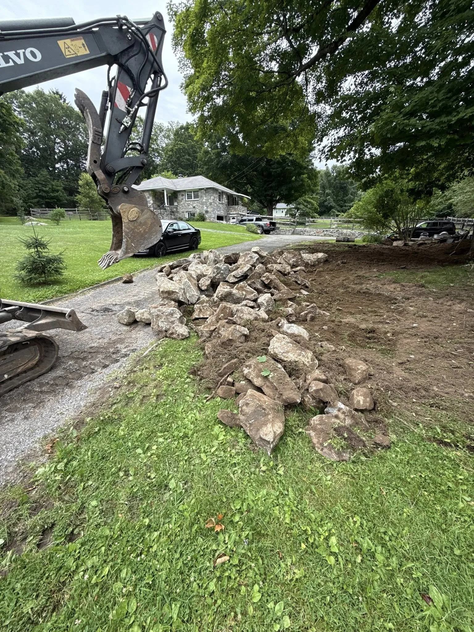 A construction site with a small excavator and a pile of rocks on a grassy area next to a dirt patch, in a residential neighborhood with houses and cars in the background.