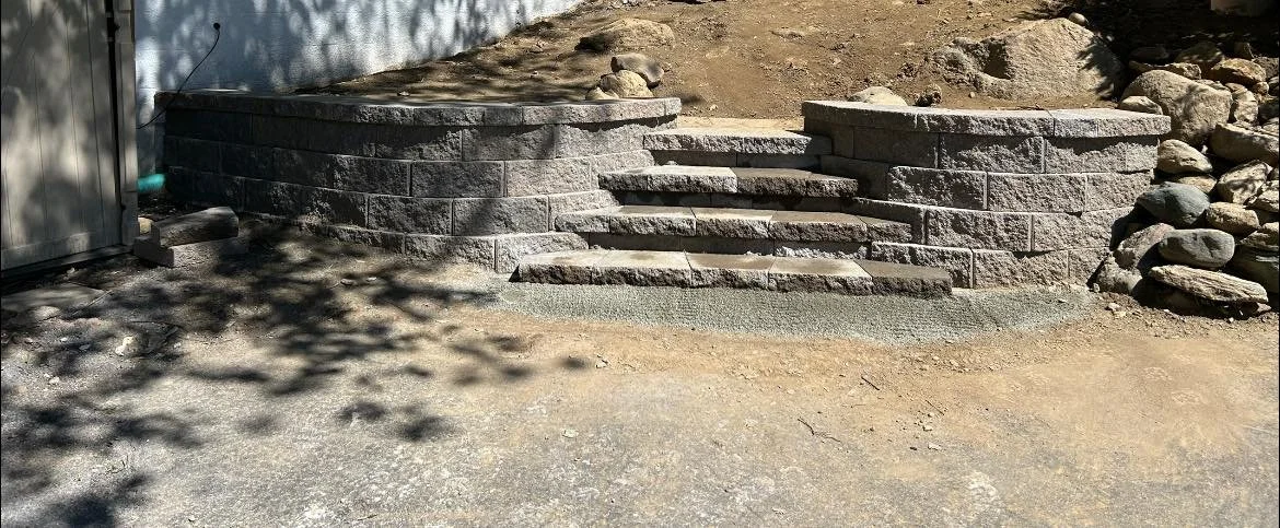 Stone staircase leading up to a raised patio area with retaining walls, outdoors, shaded by tree shadows.