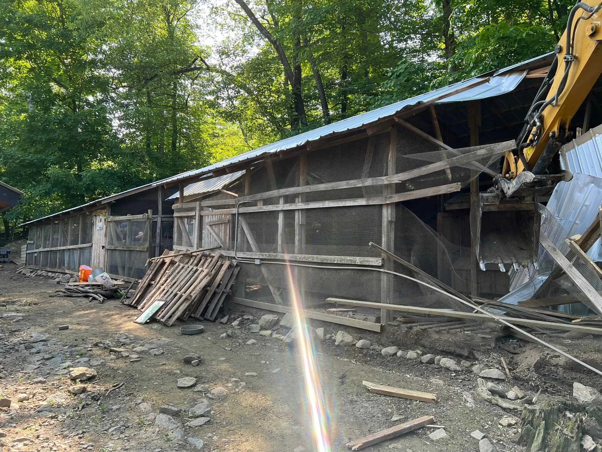 A partially demolished wooden structure with construction equipment nearby, surrounded by trees and debris on the ground.