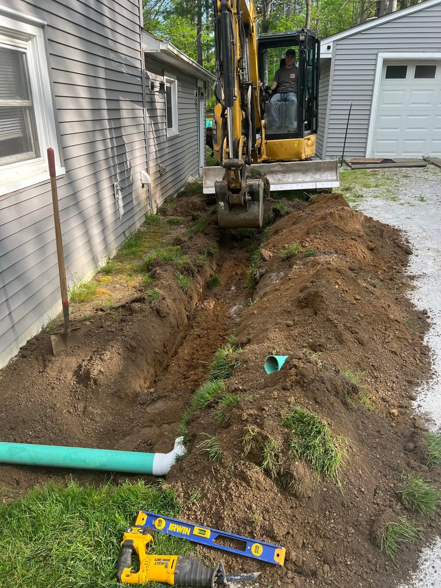 A man operating a small yellow excavator digging a trench alongside a gray house with vinyl siding. A shovel and a level tool are on the ground near the trench, and a green pipe is visible in the trench. The scene appears to be part of a plumbing or 