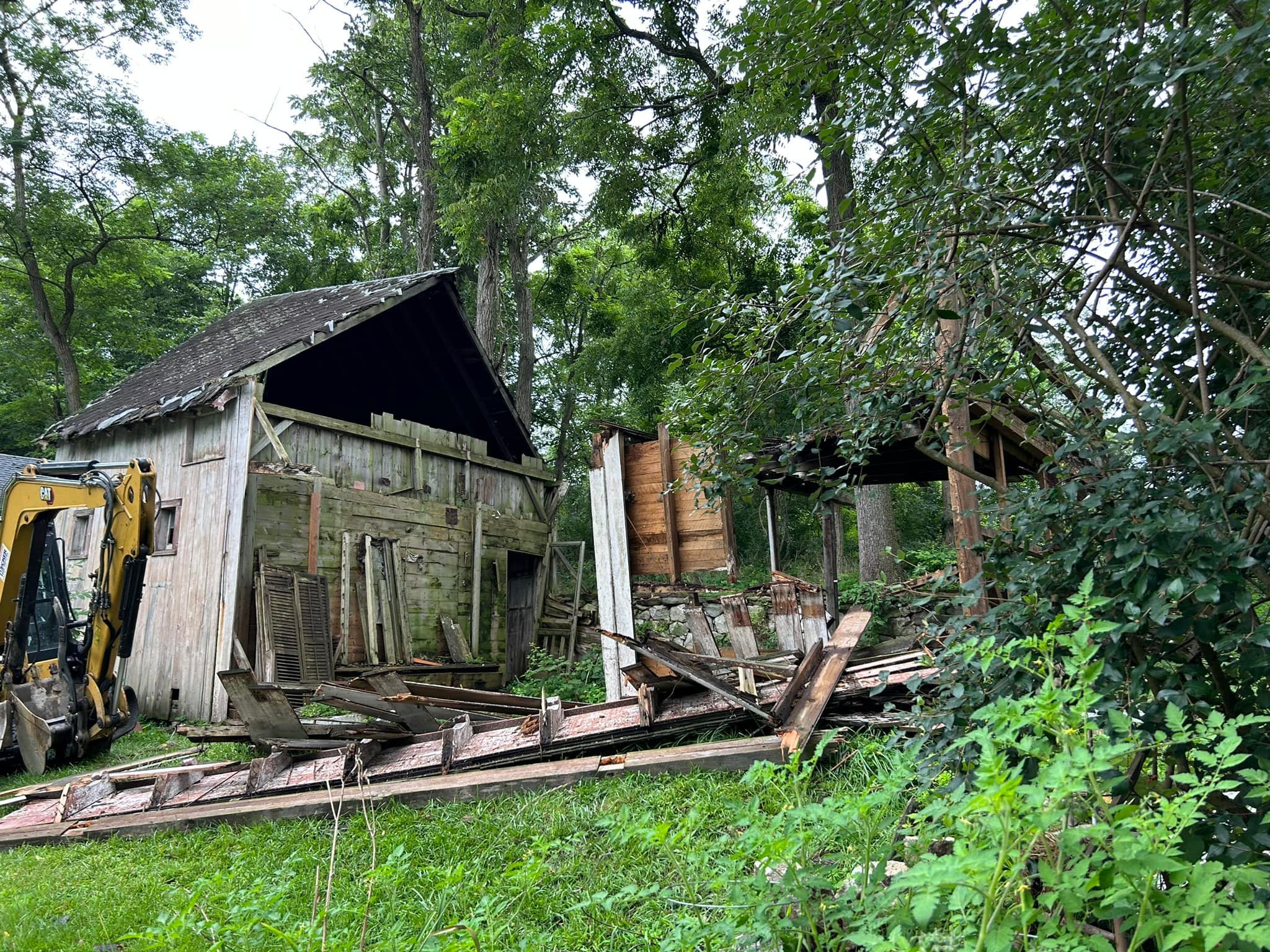 Old wooden shed partially collapsed with debris and a small excavator on the left, surrounded by green trees and grass.