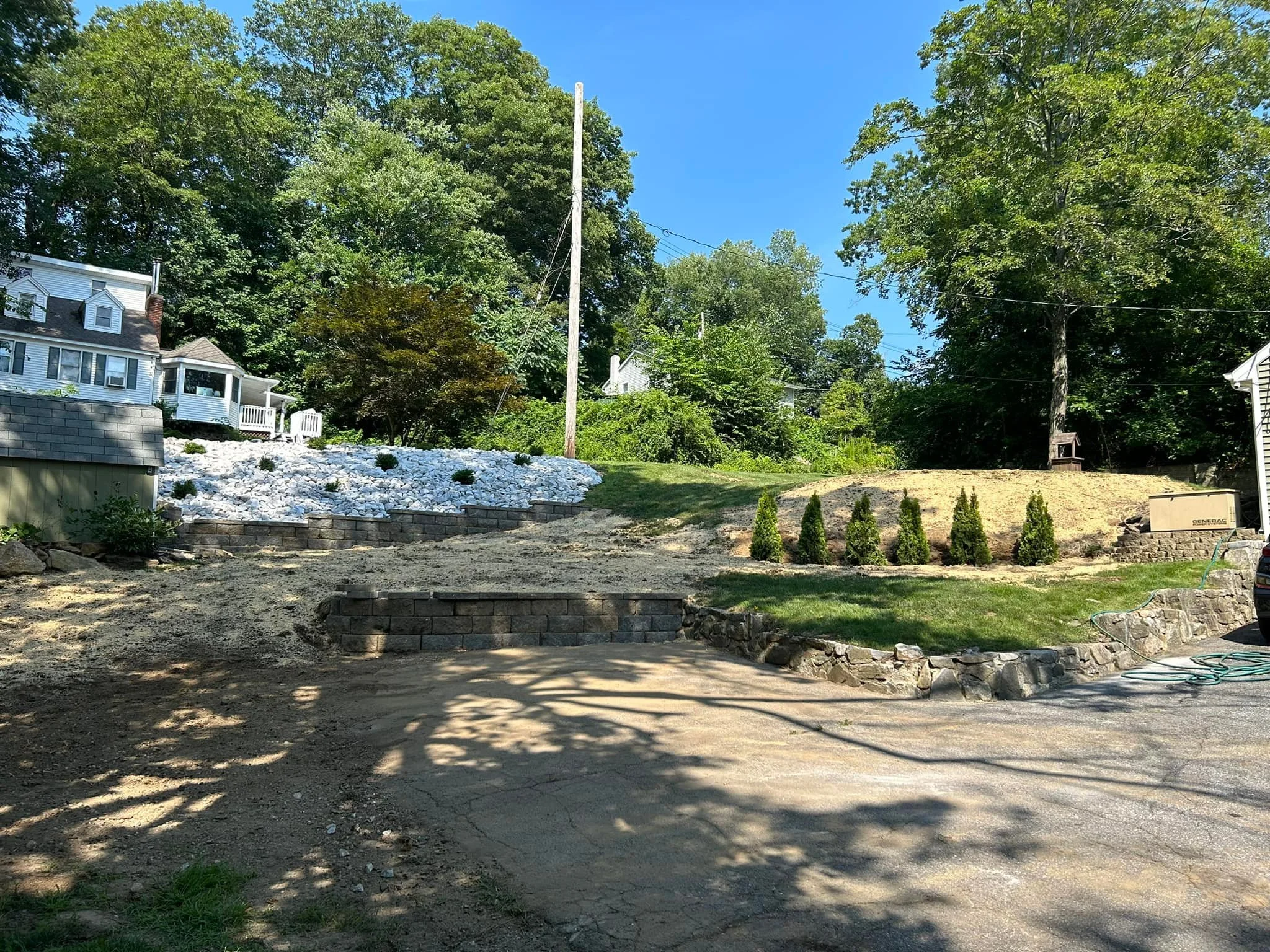 Residential backyard under landscaping with stone retaining walls, small trees, and a dirt and paved driveway, surrounded by green trees and houses.
