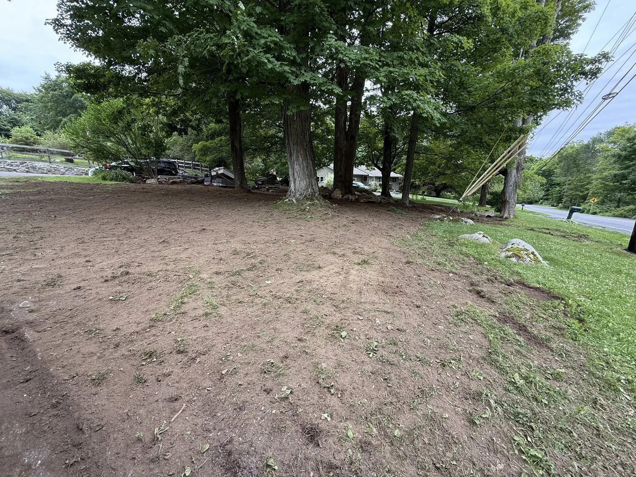 Logging equipment harvesting timber in a cleared area of forest, with stacks of cut logs and machinery in the foreground, and a rural landscape with trees and a road in the background.