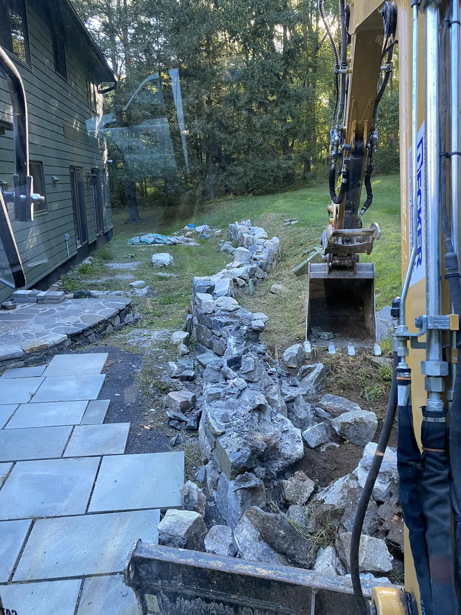Construction site with a small excavator digging a stone-lined pathway or garden, with rocks and stones piled along the edge and a house with green siding in the background.