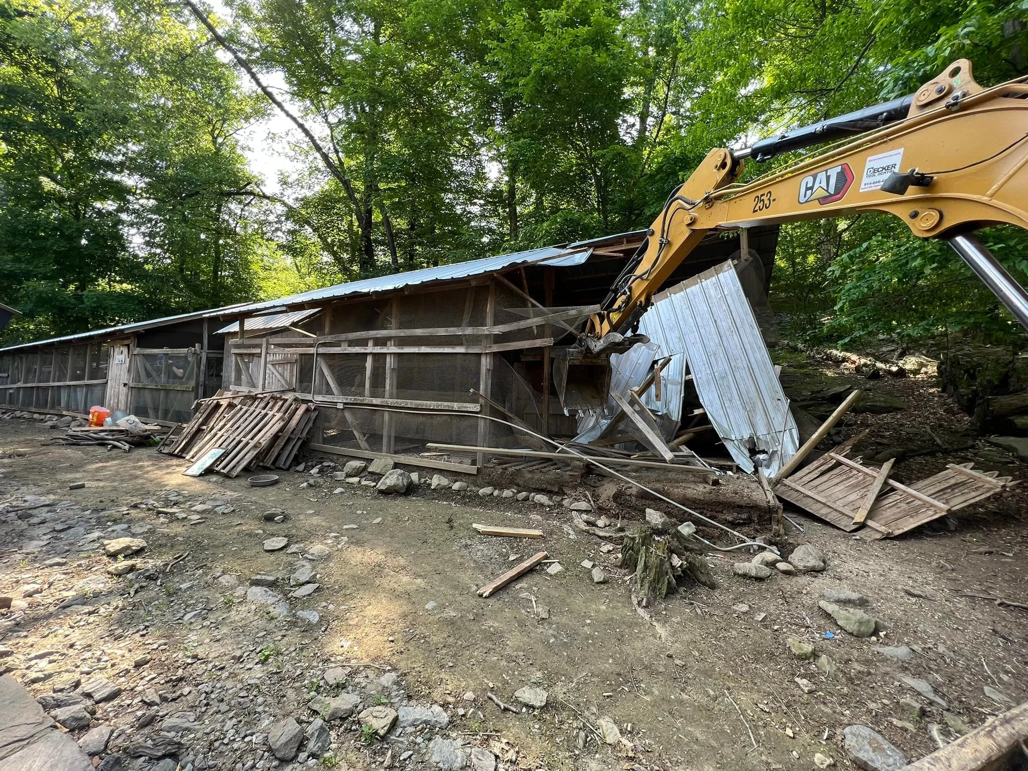 A partially collapsed wooden and metal shed being demolished by a yellow CAT excavator in a wooded area.