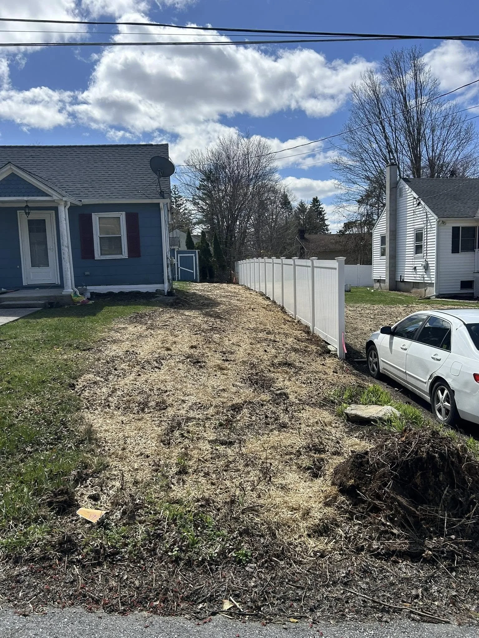 A backyard scene with two houses, a partially cleared dirt yard, a white fence, a white car parked on the street, and a partly cloudy sky.