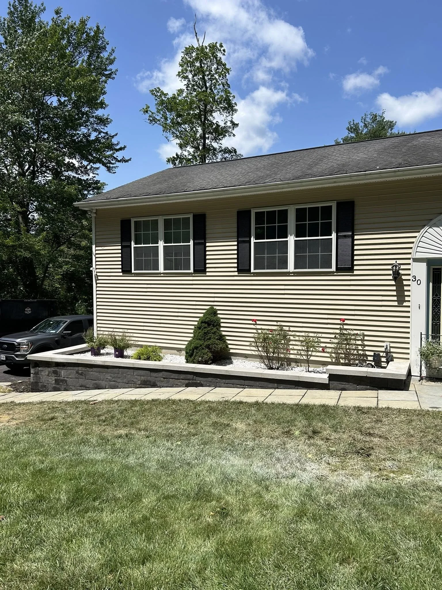 Front view of a beige house with black shutters, a small garden bed with shrubs and flowers, a sidewalk, and a parked vehicle on the driveway, under a partly cloudy sky.