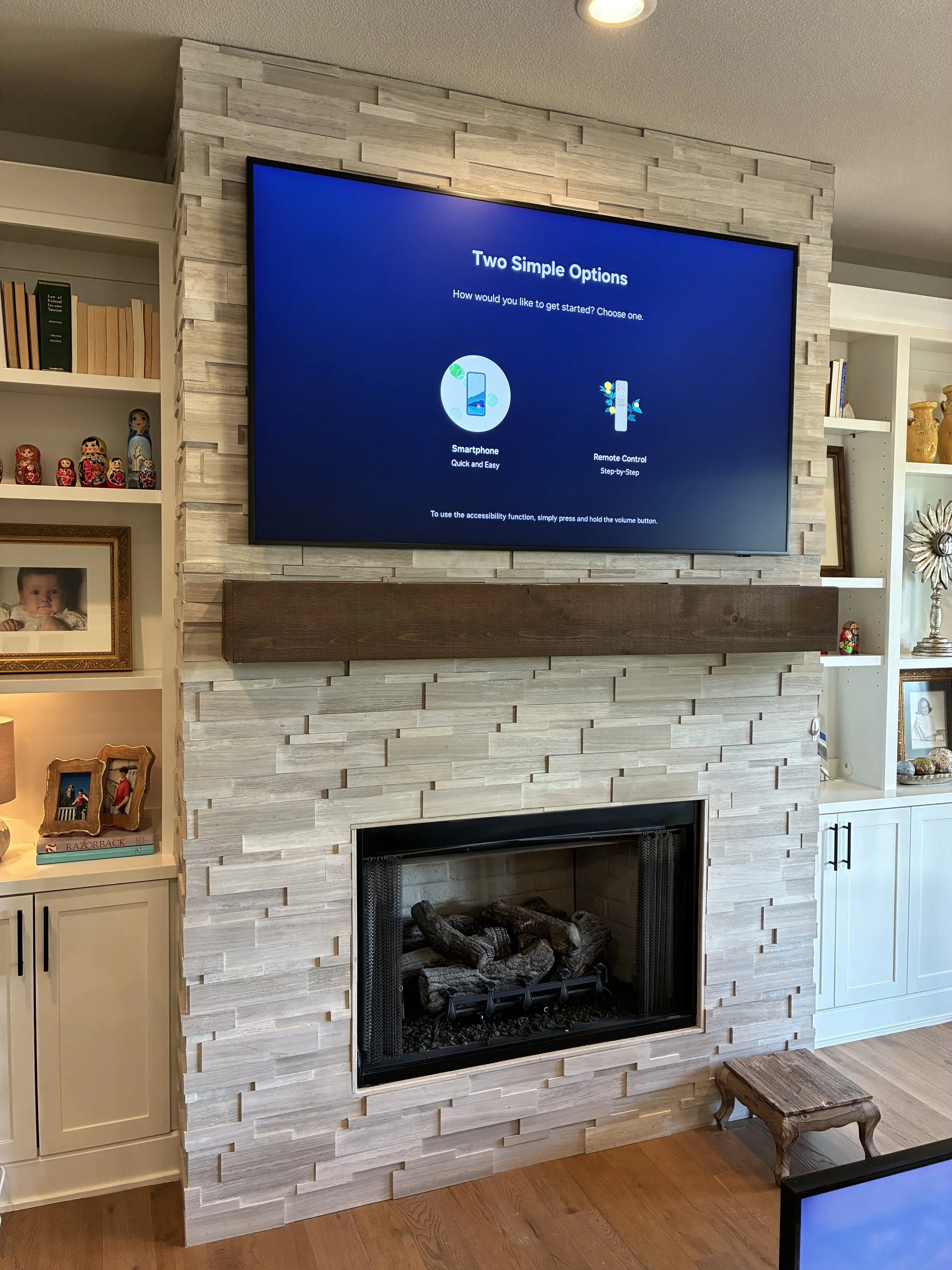 Living room fireplace with a stone surround, a mounted TV displaying two options for device setup, and decorations including photos, books, and figurines.