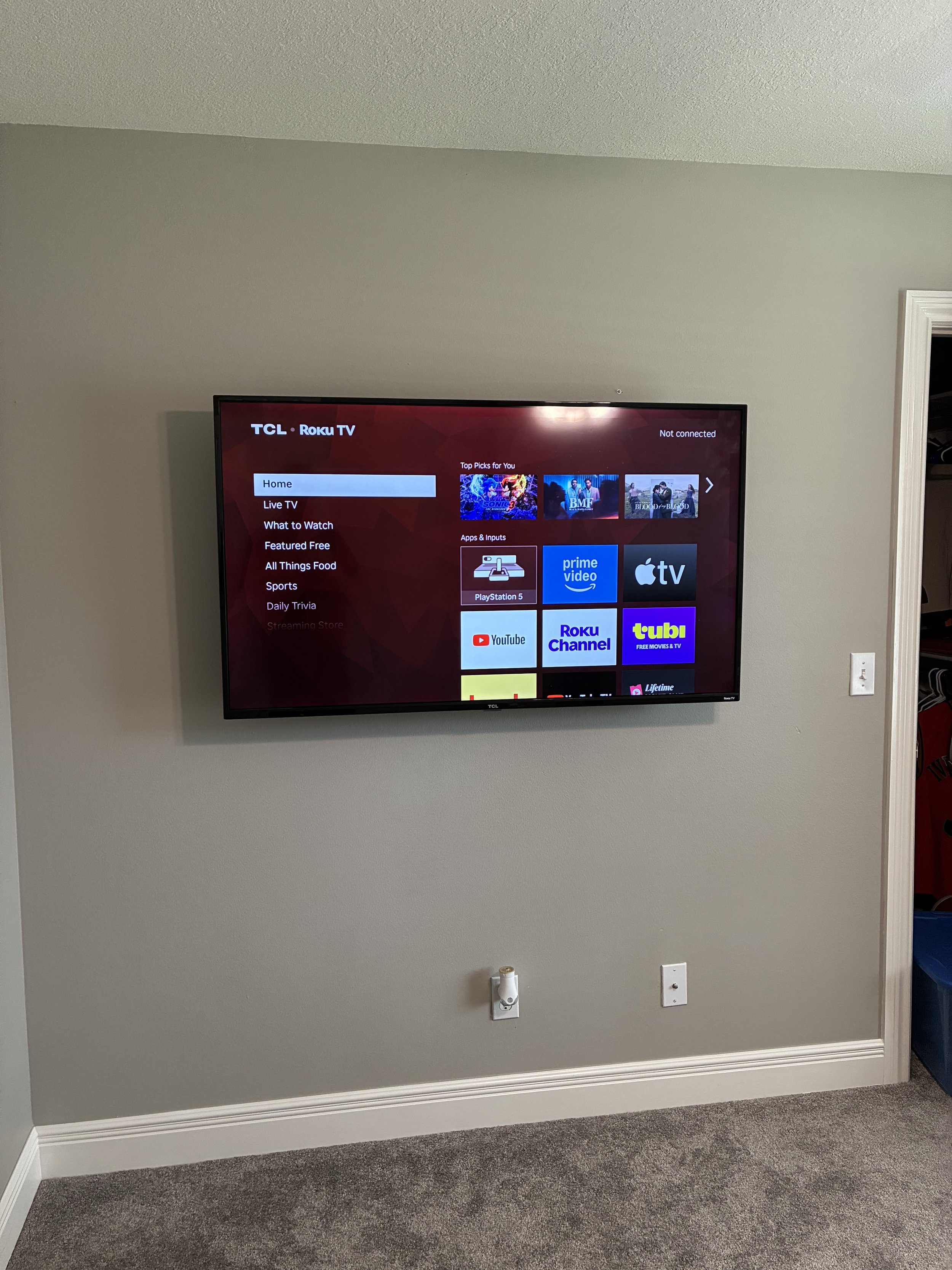 Flat-screen TV mounted on a beige wall, displaying streaming options and apps like YouTube, Roku Channel, and Prime Video.