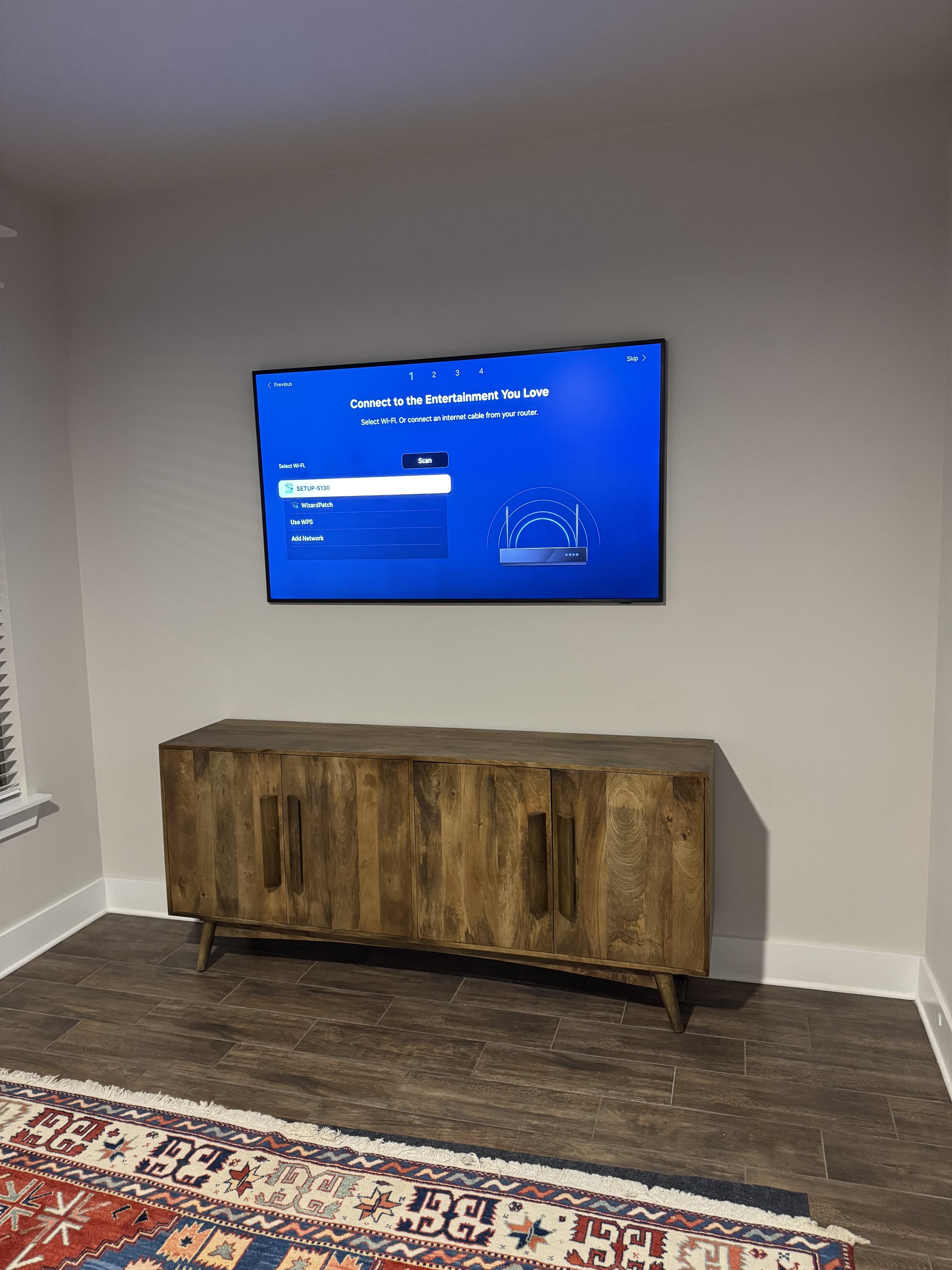 A living room with a wall-mounted flat-screen TV displaying a setup menu, a wooden TV stand, a colorful patterned rug, and hardwood flooring.