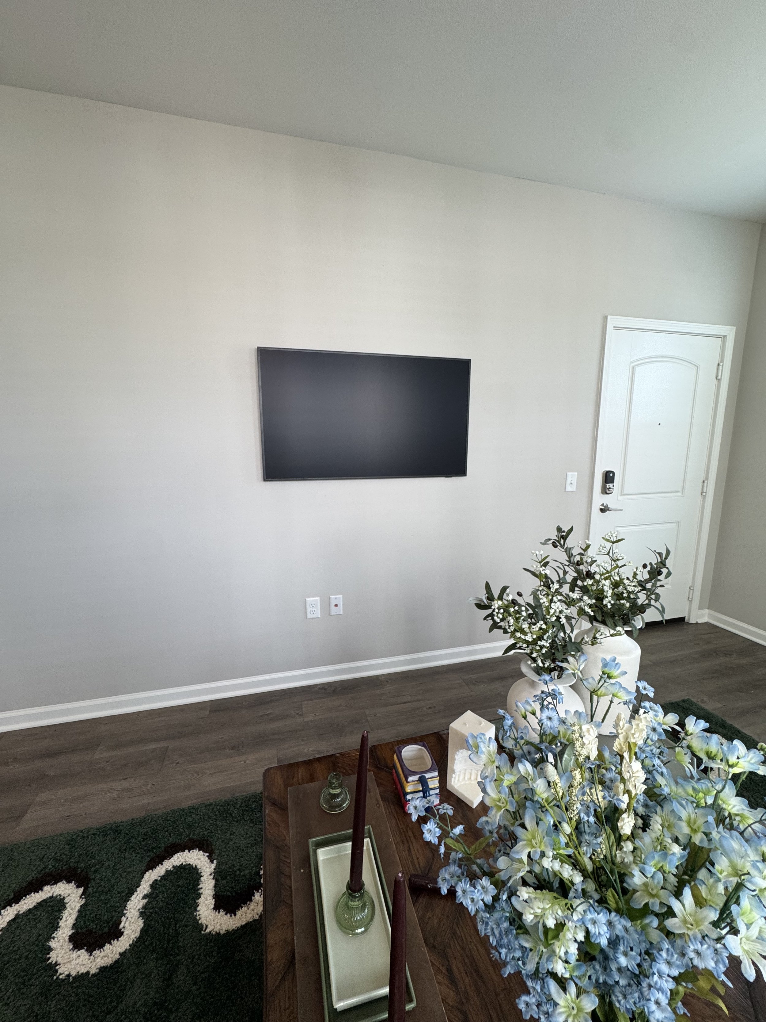 Living room with a mounted flat-screen TV on a white wall, a wooden table with decorative candles and a tray, a large floral arrangement, and a closed front door.