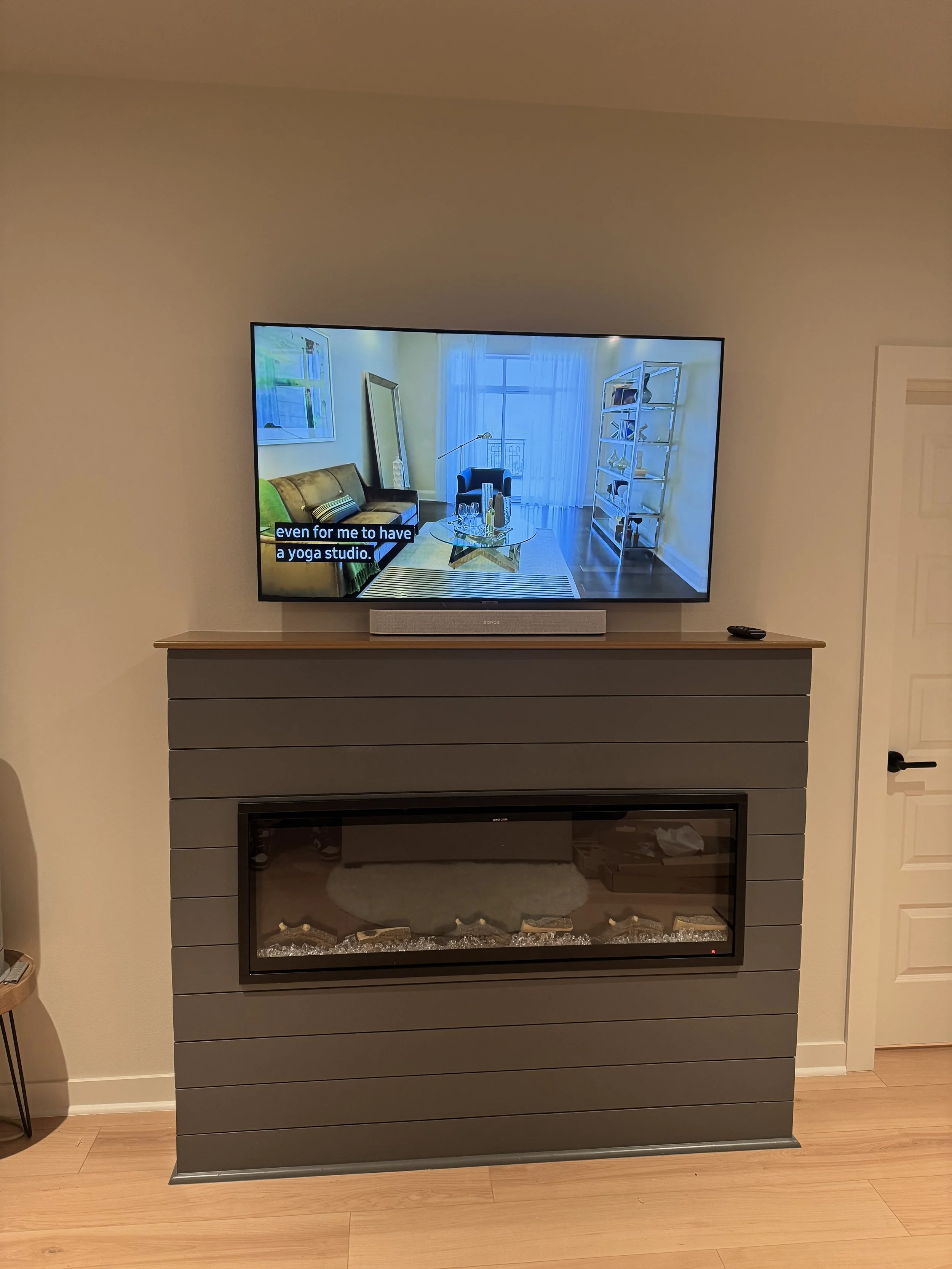 Modern living room with a wall-mounted TV above a gray fireplace, displaying a room with a sofa, armchair, bookshelf, and large window with curtains.