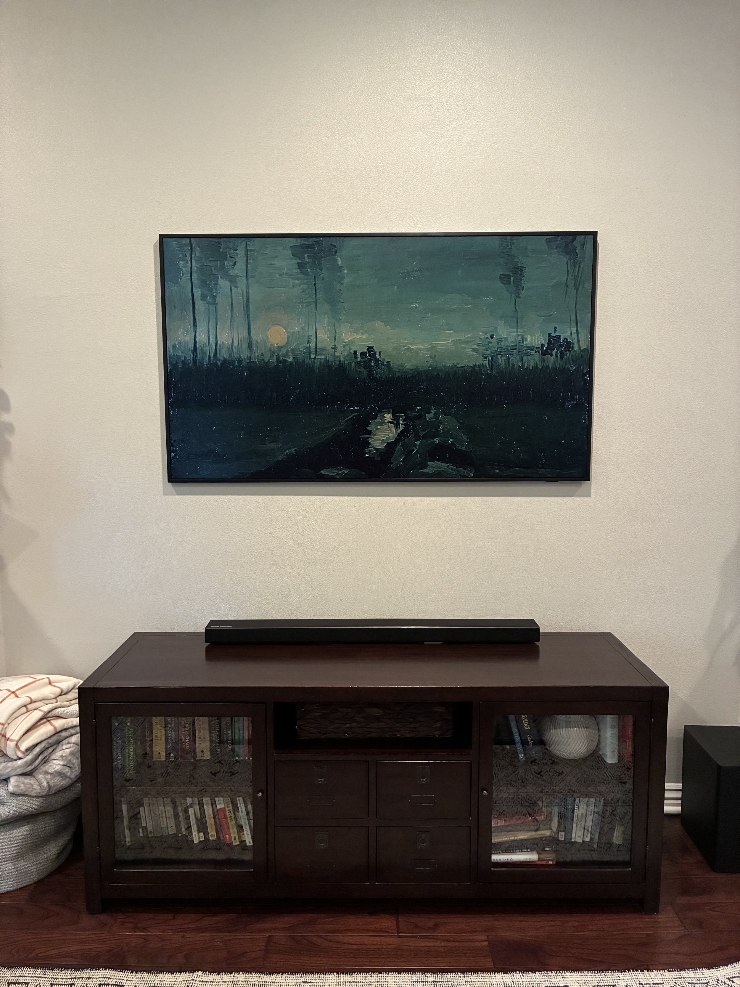 A dark wooden media console with glass doors and drawers, topped with a long black soundbar, against a white wall, with a landscape painting of a blue-green sunset or sunrise scene hanging above.