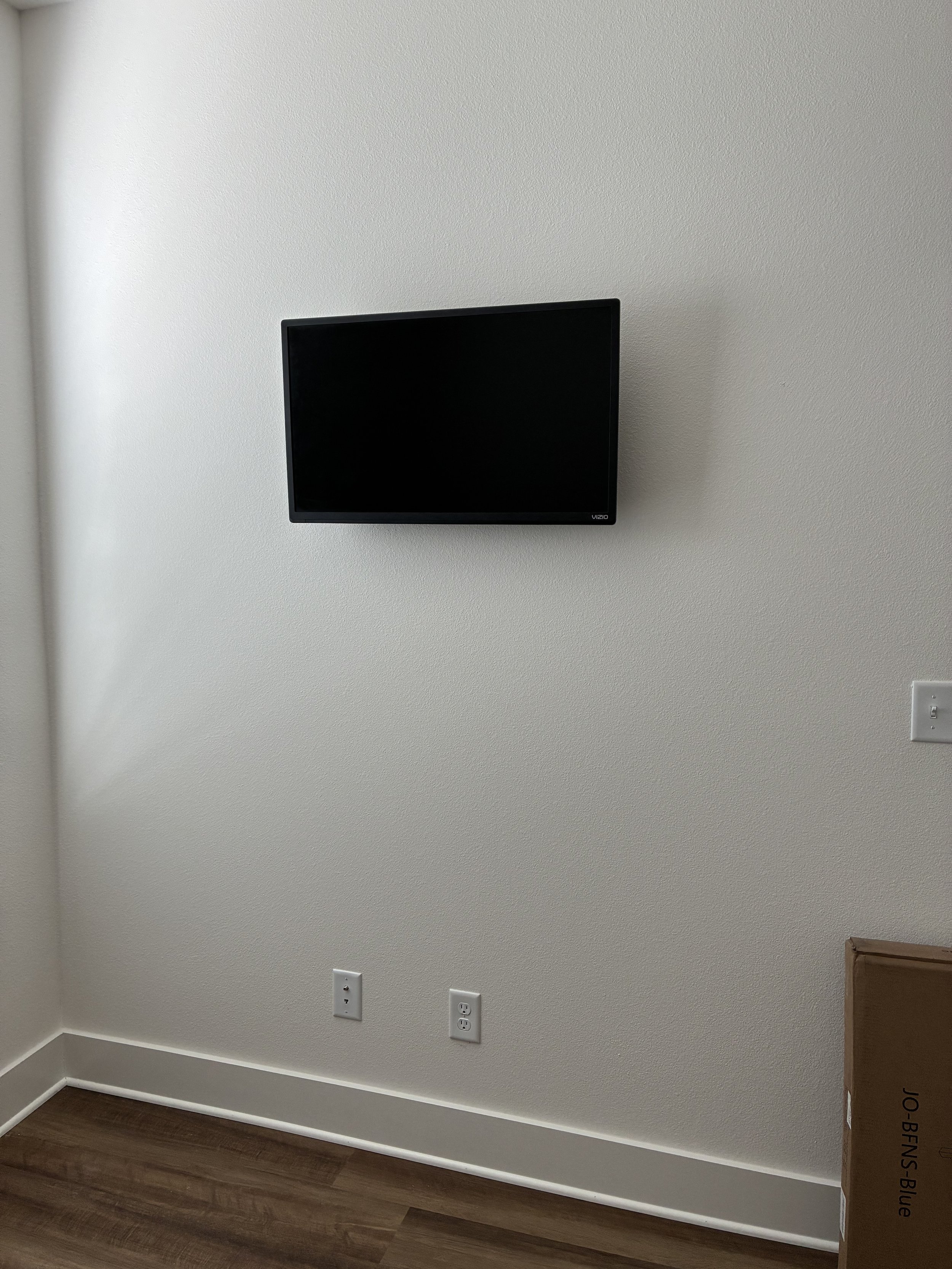 A small flat-screen TV mounted on a white wall in a room with wood flooring. There are three power outlets near the floor and a cardboard box labeled 'JO-BENS Blue' on the right side of the image.