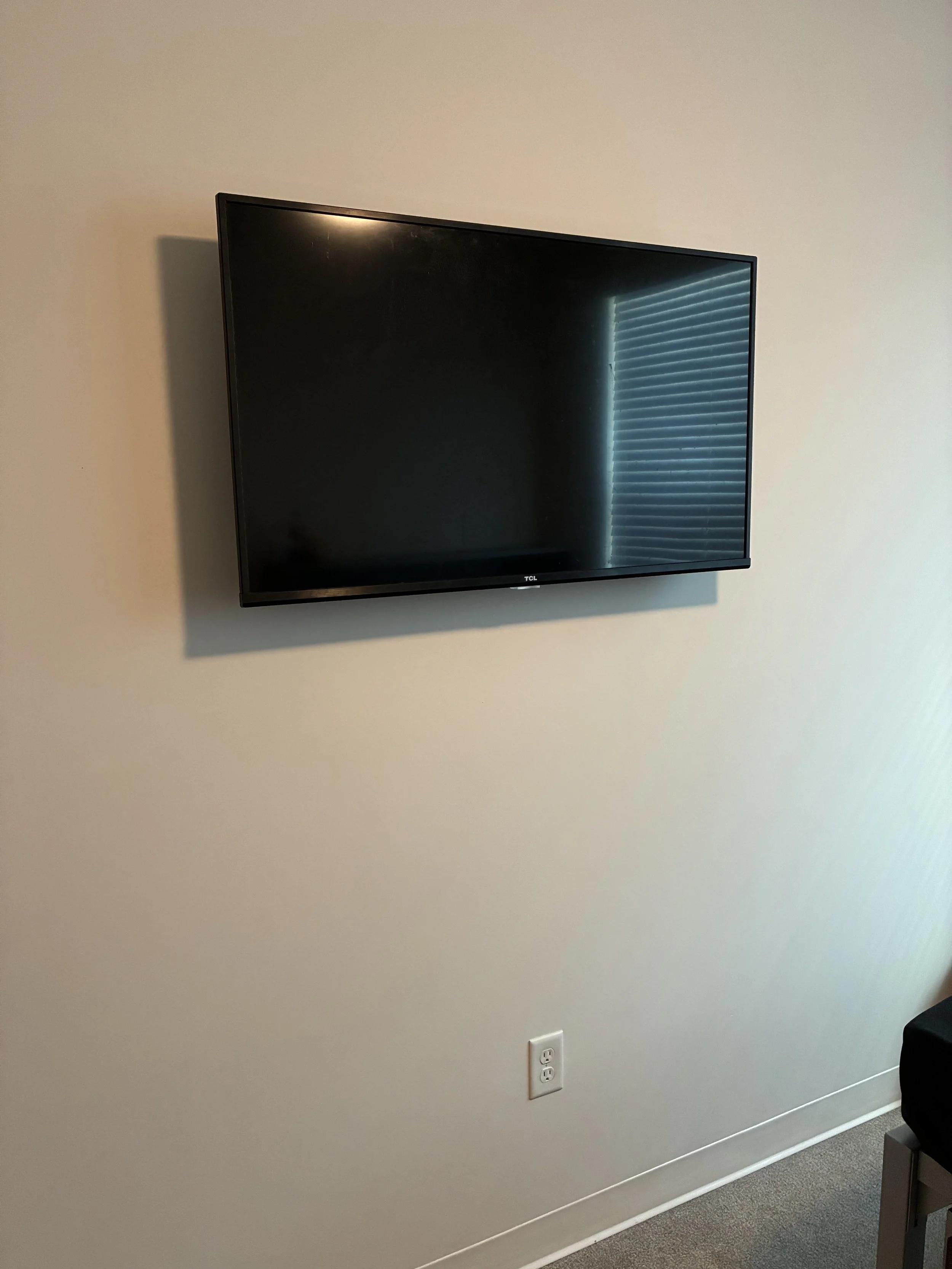 Flat-screen television mounted on a beige wall next to a window with blinds, a power outlet below, and a piece of furniture partially visible on the right.