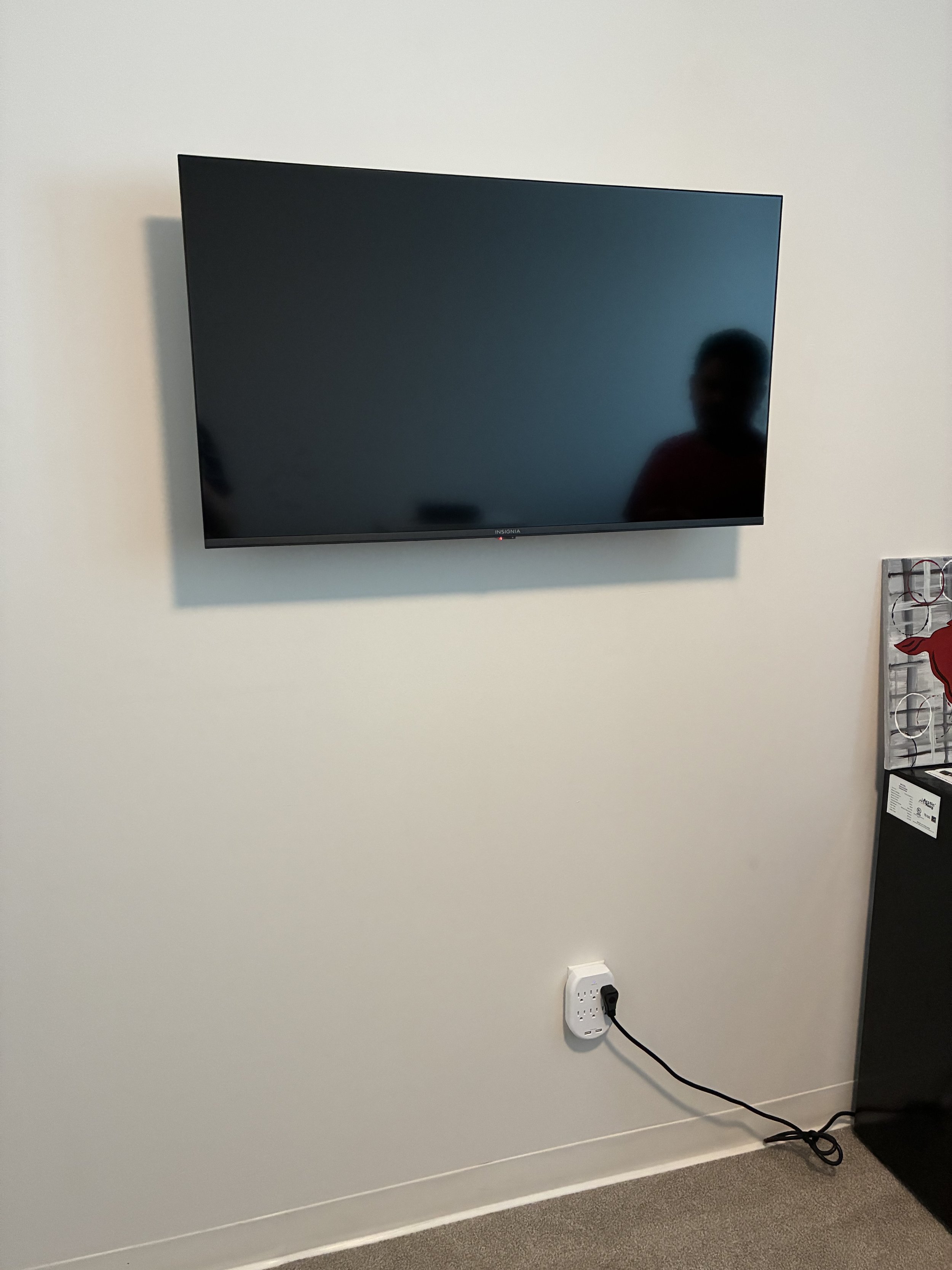 Flat-screen television mounted on a white wall, with a reflection of a person wearing a red shirt.