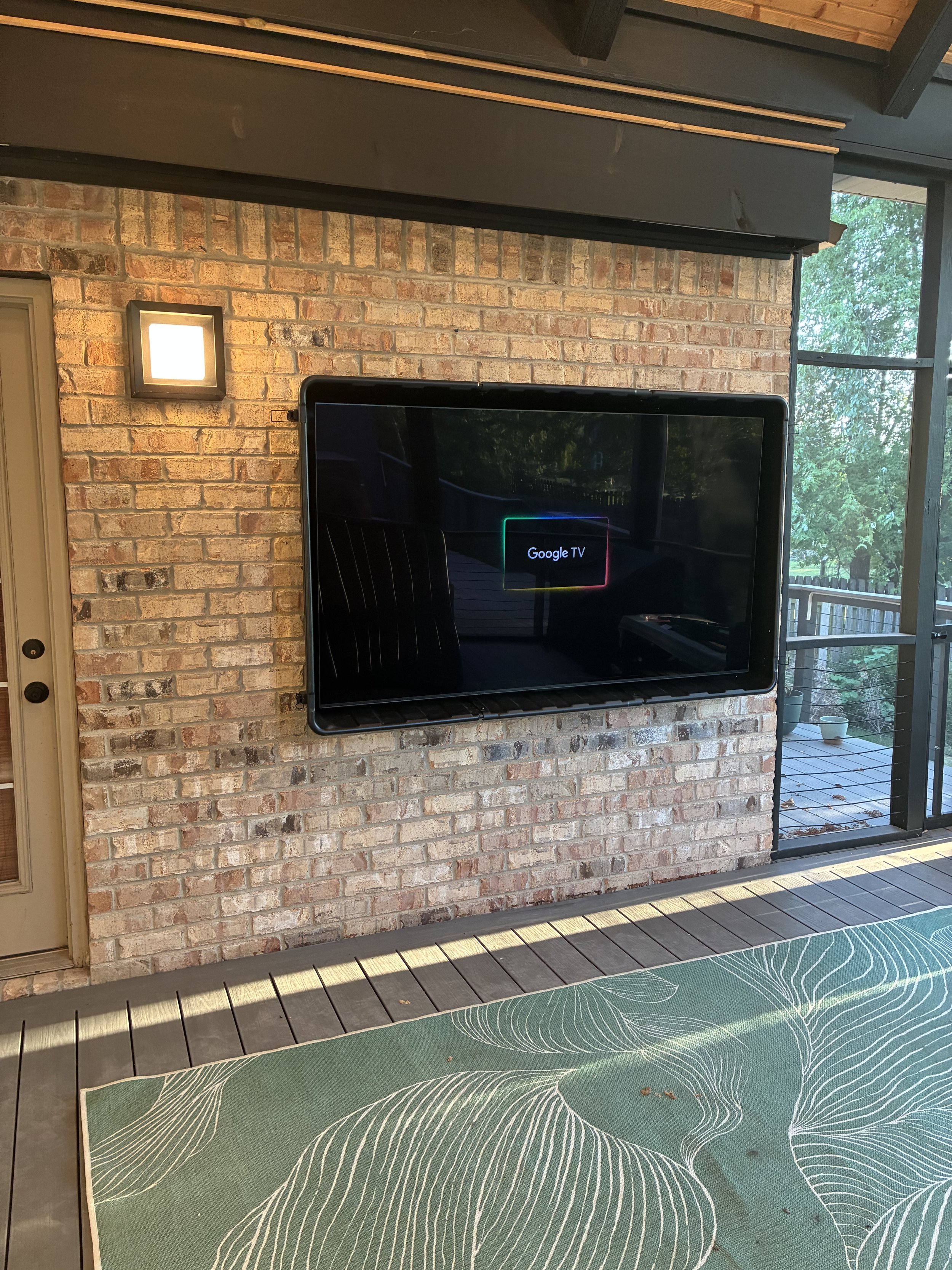 A flat-screen TV mounted on a brick wall displaying the Google TV logo. To the right, there's a window with a view of a balcony and trees outside. The foreground shows a green rug with a leaf pattern on a wooden deck.