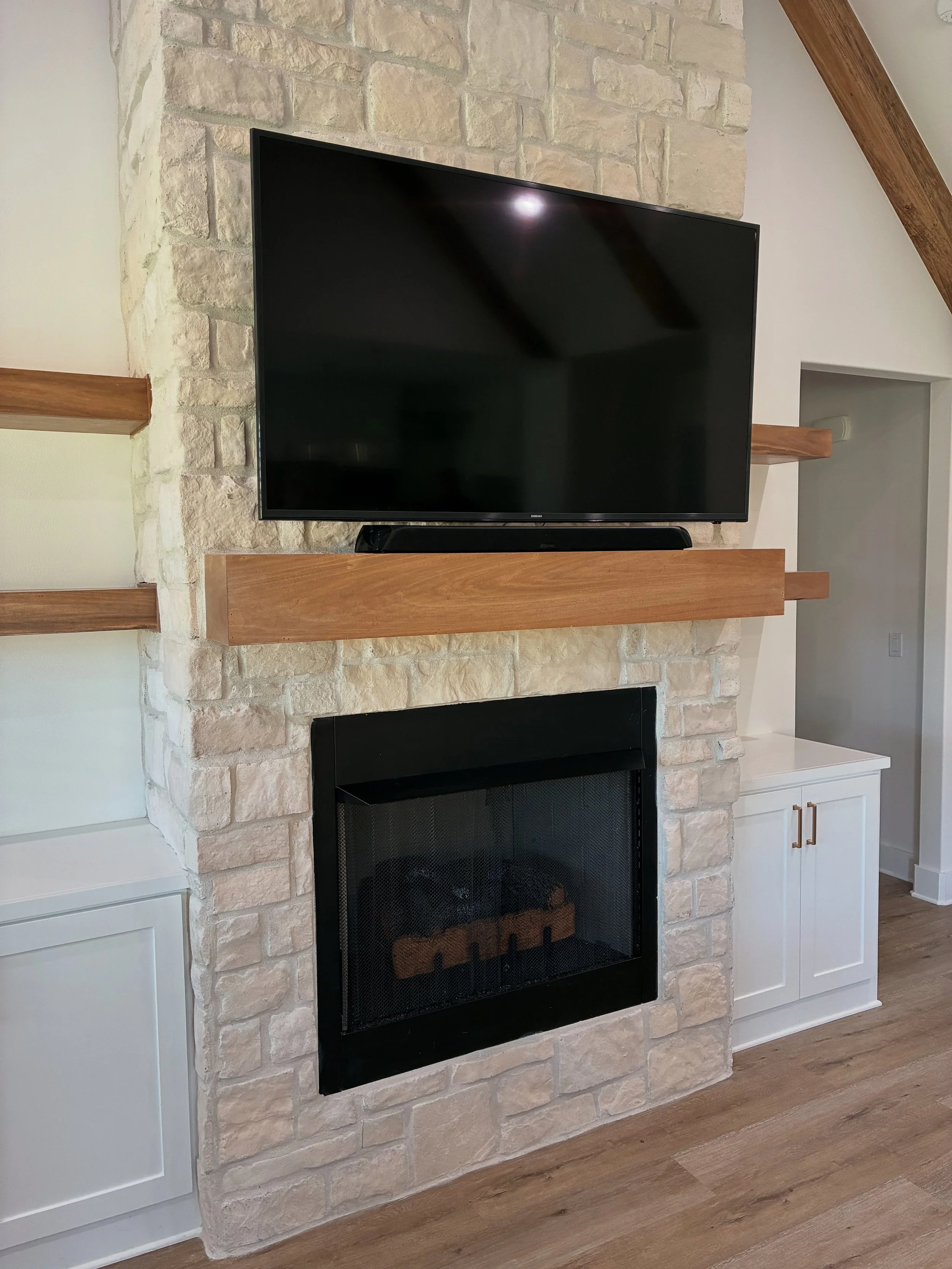 Living room fireplace with a mounted flat-screen TV above, cream-colored brick surround, black firebox, wooden mantel, and white cabinets on both sides. Wooden beams visible on the ceiling.