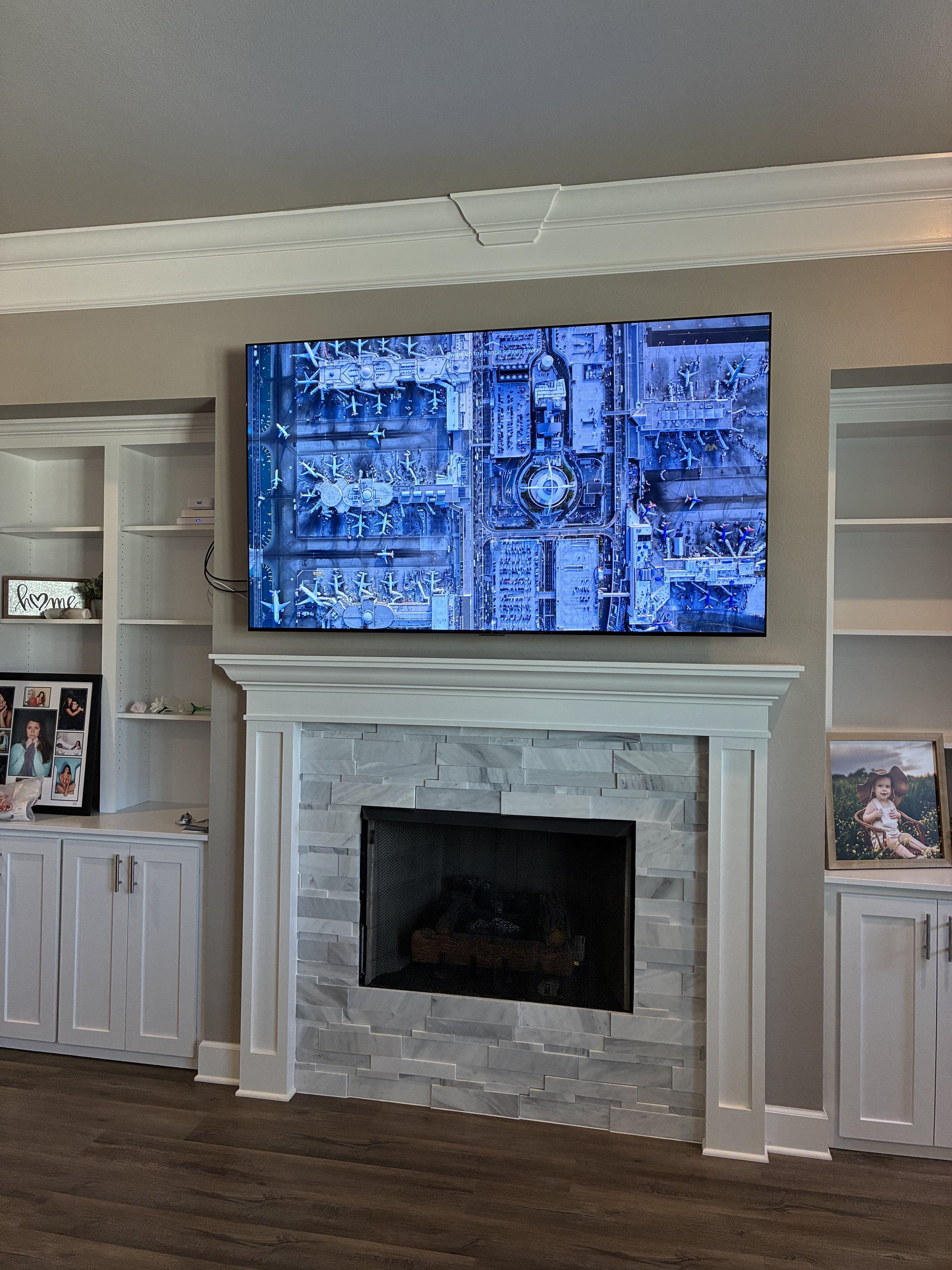 Living room with a wall-mounted television above a white fireplace, white built-in shelves on either side, and family photos displayed on the left shelf and a framed photo on the right shelf. The TV screen shows an aerial view of an amusement park wi