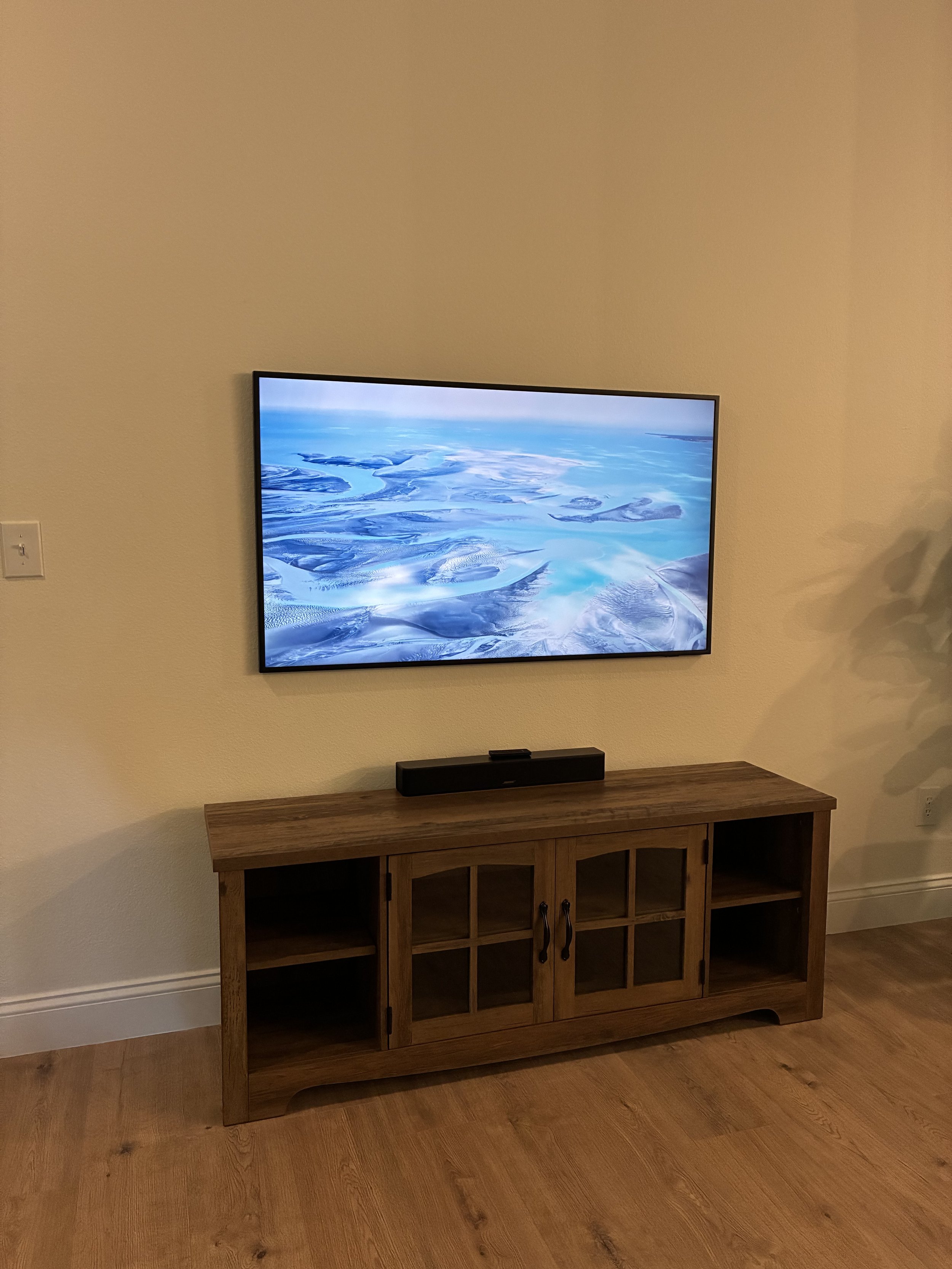A flat-screen TV displaying an aerial view of icy and snowy landscapes, mounted on a white wall above a wooden TV stand with glass-paneled doors and open shelves, in a room with wooden flooring.