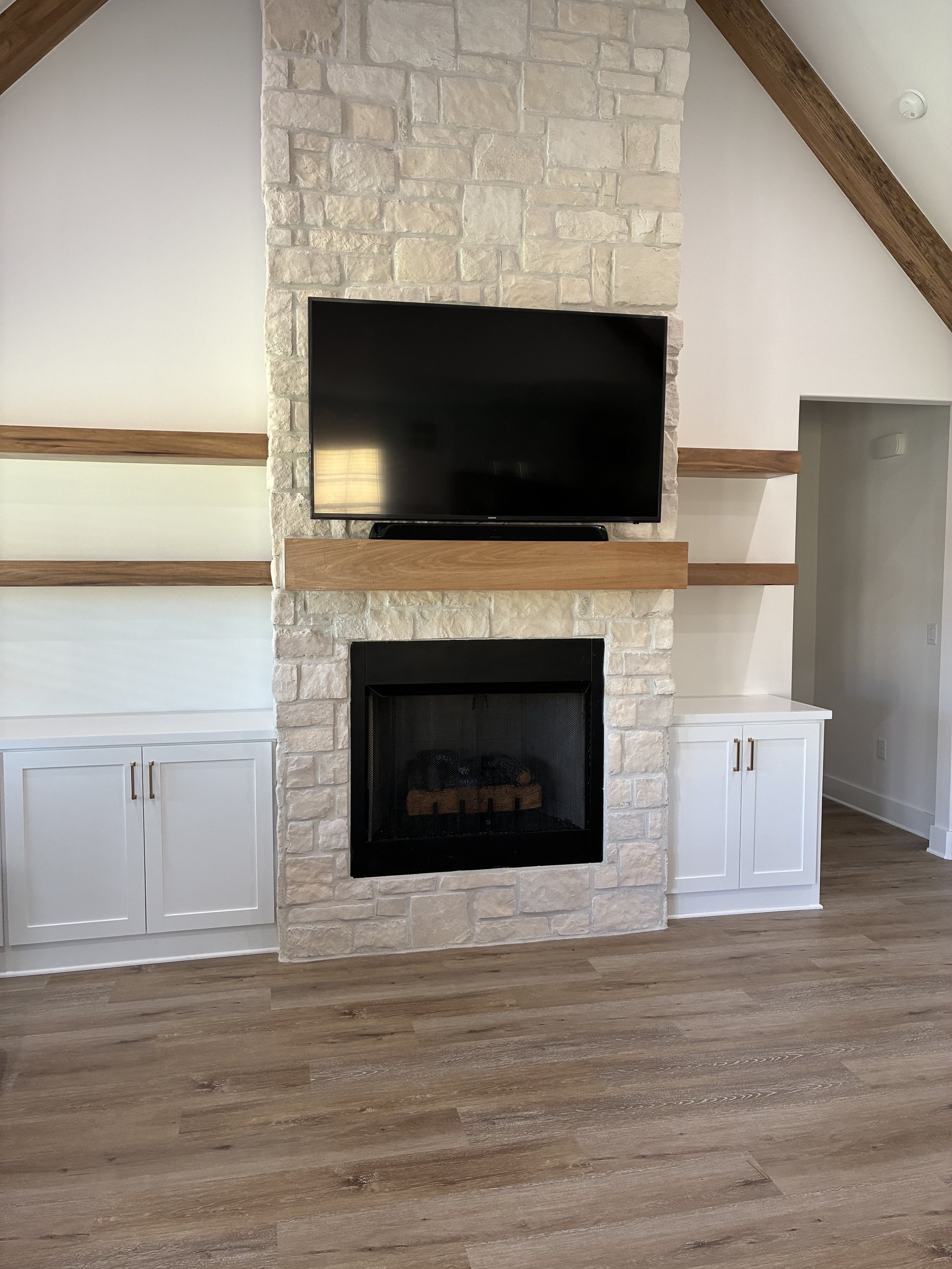 Living room with a stone fireplace, mounted flat-screen TV, white built-in cabinets, and wooden trim and shelving.