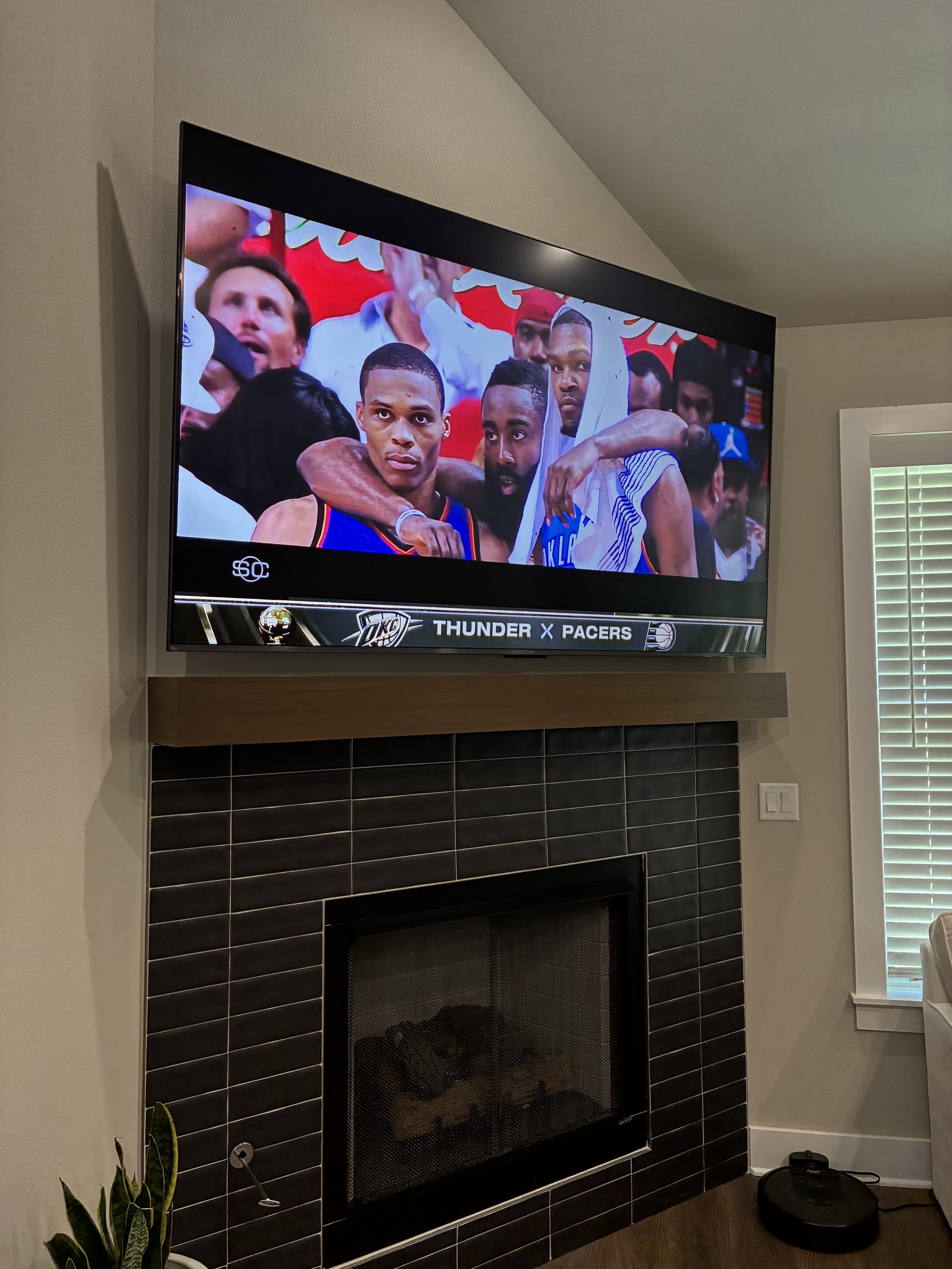 Living room with a flat-screen TV displaying a basketball game, mounted above a fireplace with black tiles, and a plant near the fireplace.