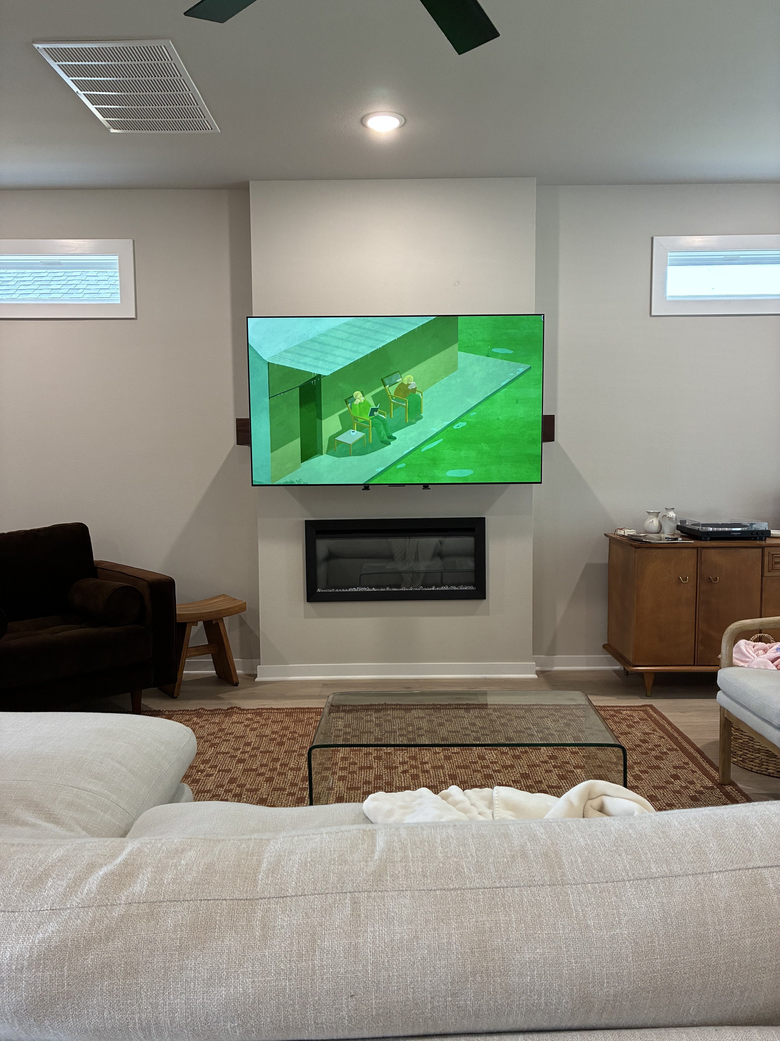 Living room with beige sofa, glass coffee table, fireplace, wooden cabinet with decorative items, and a wall-mounted television showing an animated scene. Small windows near the ceiling and a ceiling fan with lights.